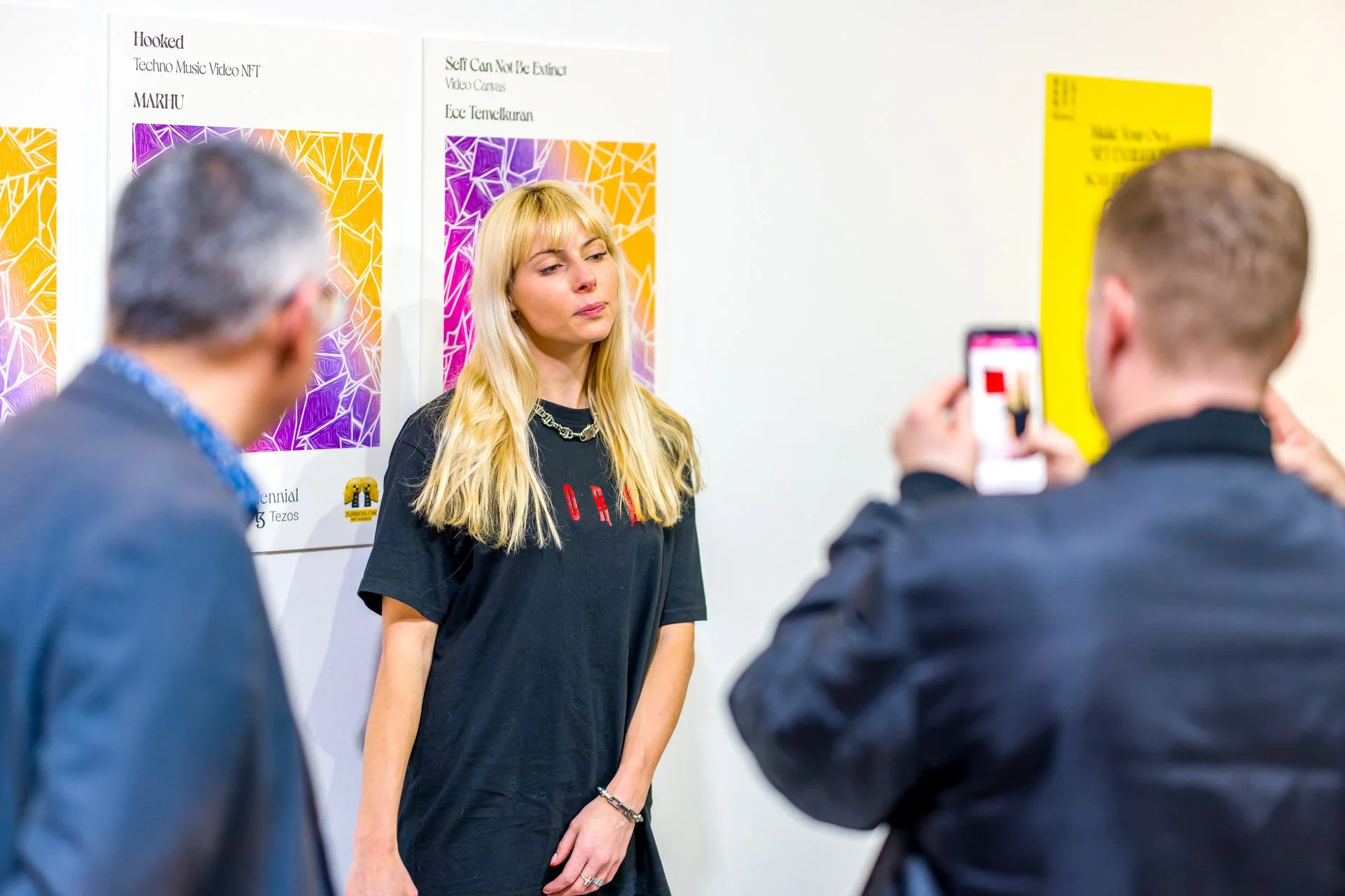 Two people are taking photos of a blonde woman standing in front of colourful posters on a white wall.