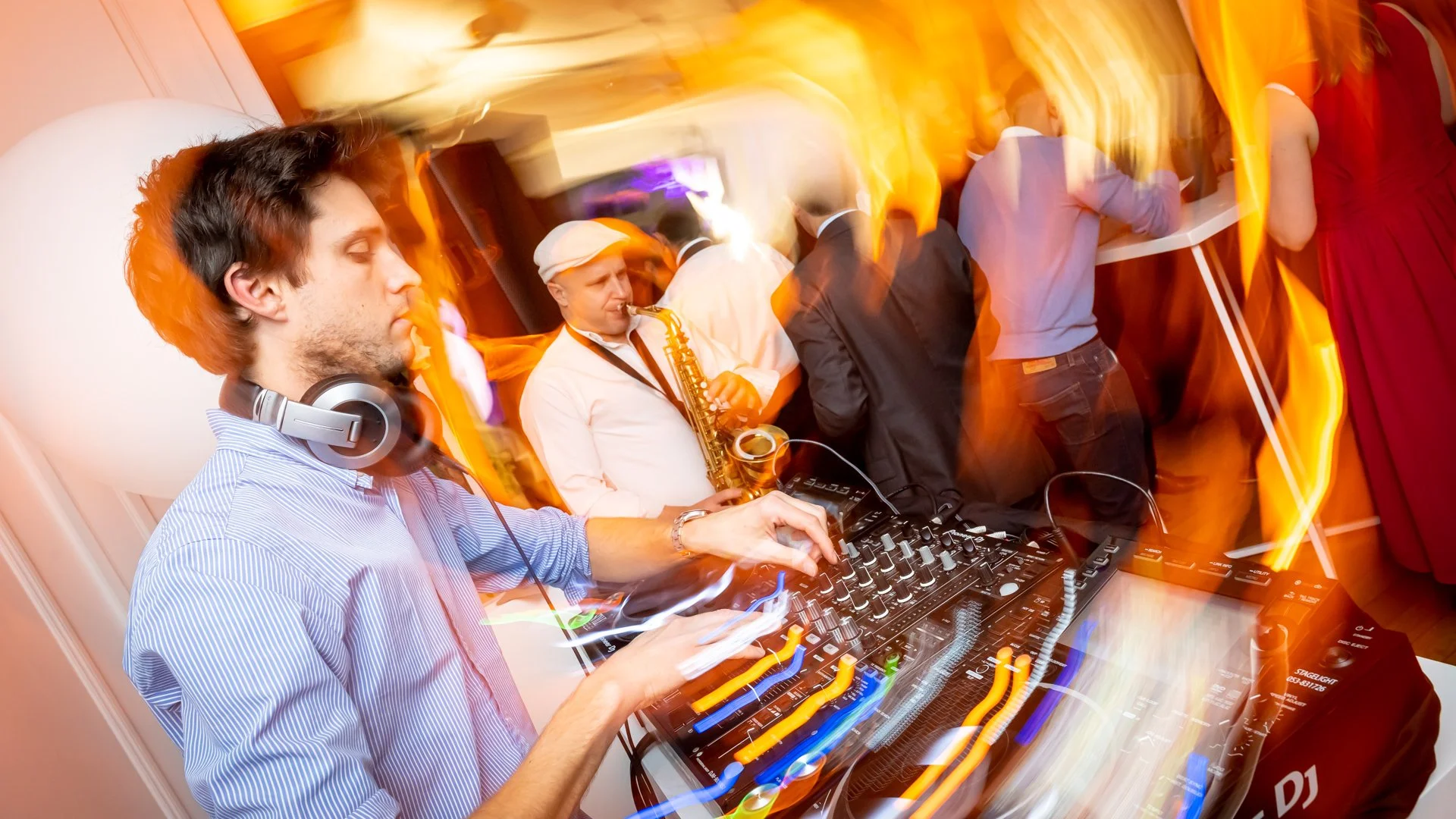A DJ mixing on turntables, wearing headphones, whilst a saxophonist plays music in the background. Coloured lights create a festive atmosphere and people are dancing around.