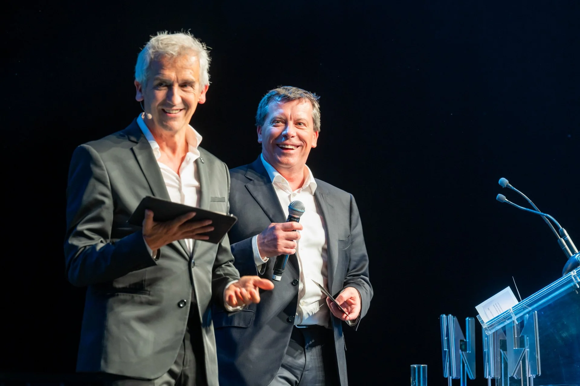 Bruno Coppens and Michel Daerden on stage during a ceremony, one holding a document and the other a glass.
