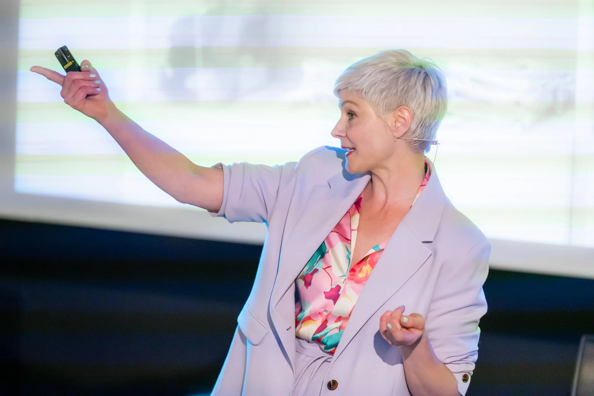 A female speaker with short hair is presenting projected material, her arm outstretched and a remote control in her hand.