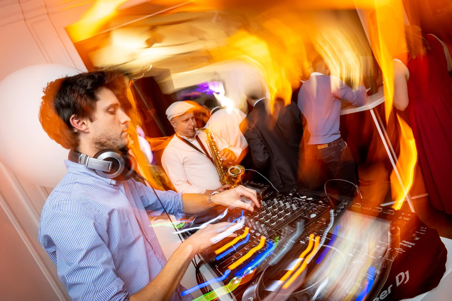 A DJ, with headphones slung round his neck, is operating his mixing desk at a corporate party, bathed in orange lighting.