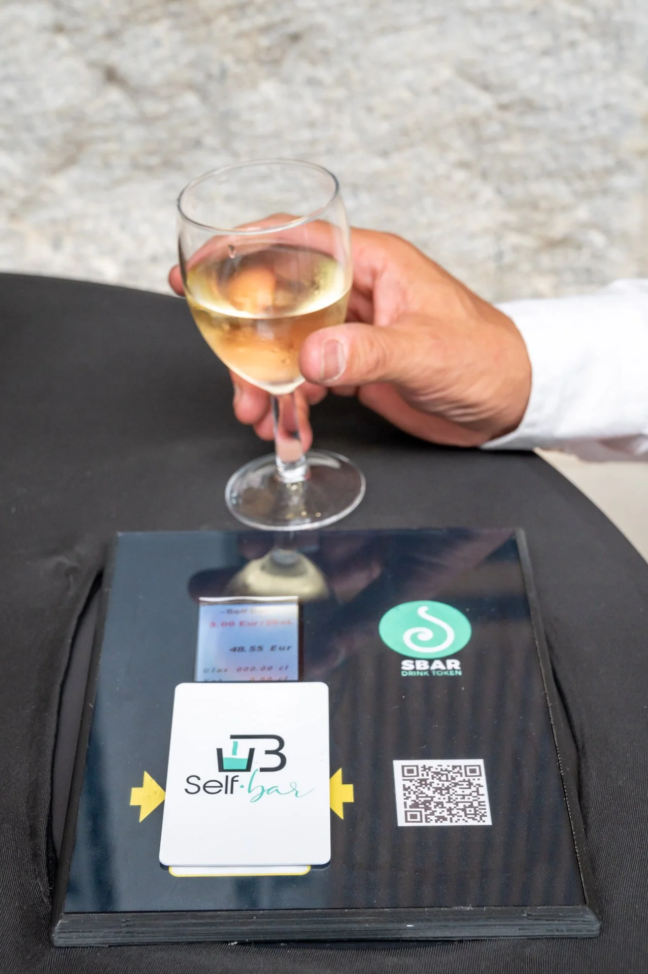 A hand is holding a glass above a self-service terminal fitted with a payment reader and a QR code.