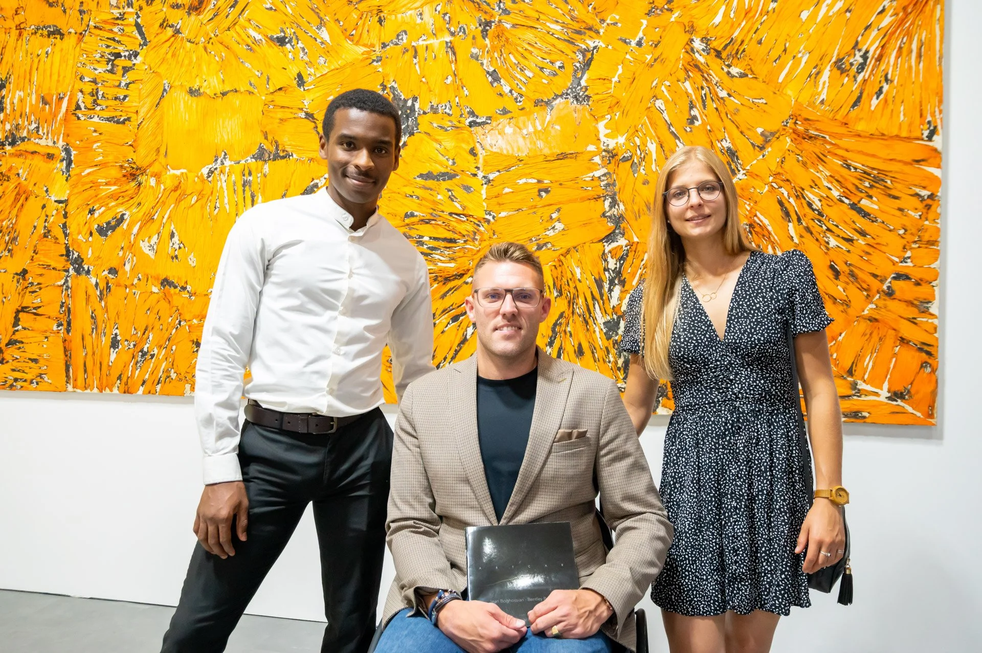 Nigel Bailly, in a wheelchair, poses between two visitors in front of a large yellow painting at the BXL ART NFT Gallery.