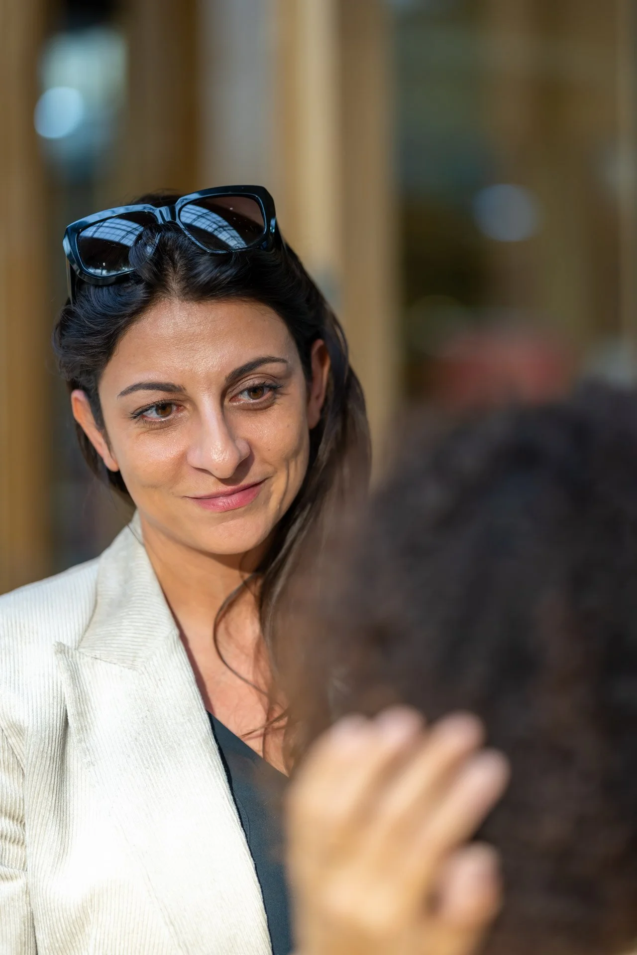 Mehri Ghaleghovand, wearing a light-coloured jacket and sunglasses perched on her head, is chatting to someone in the foreground at the BXL ART NFT Gallery.