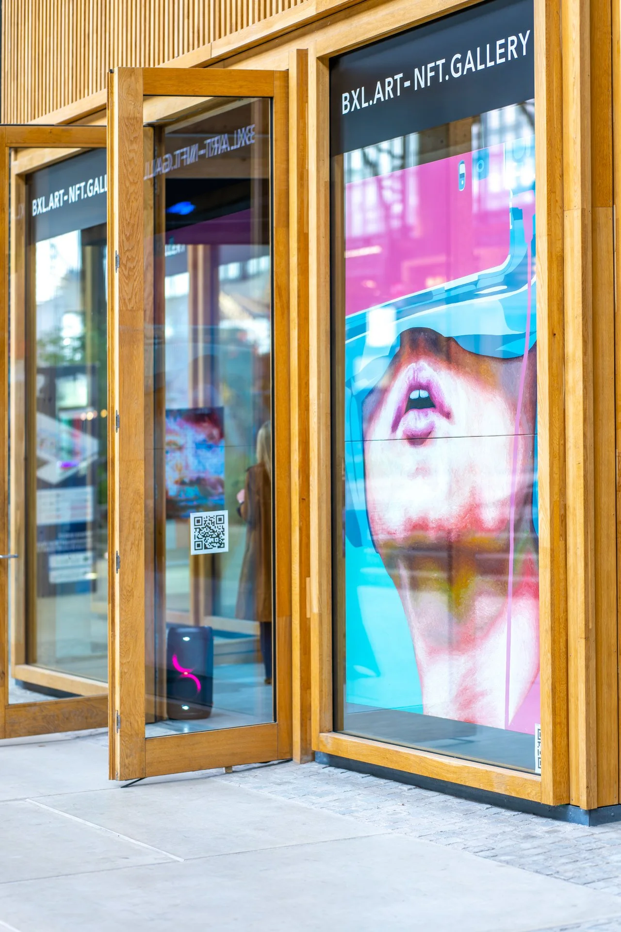 Entrée d'une galerie NFT avec une grande affiche d'art numérique montrant un visage portant un casque de réalité virtuelle, entourée de portes en bois, avec un QR code visible.