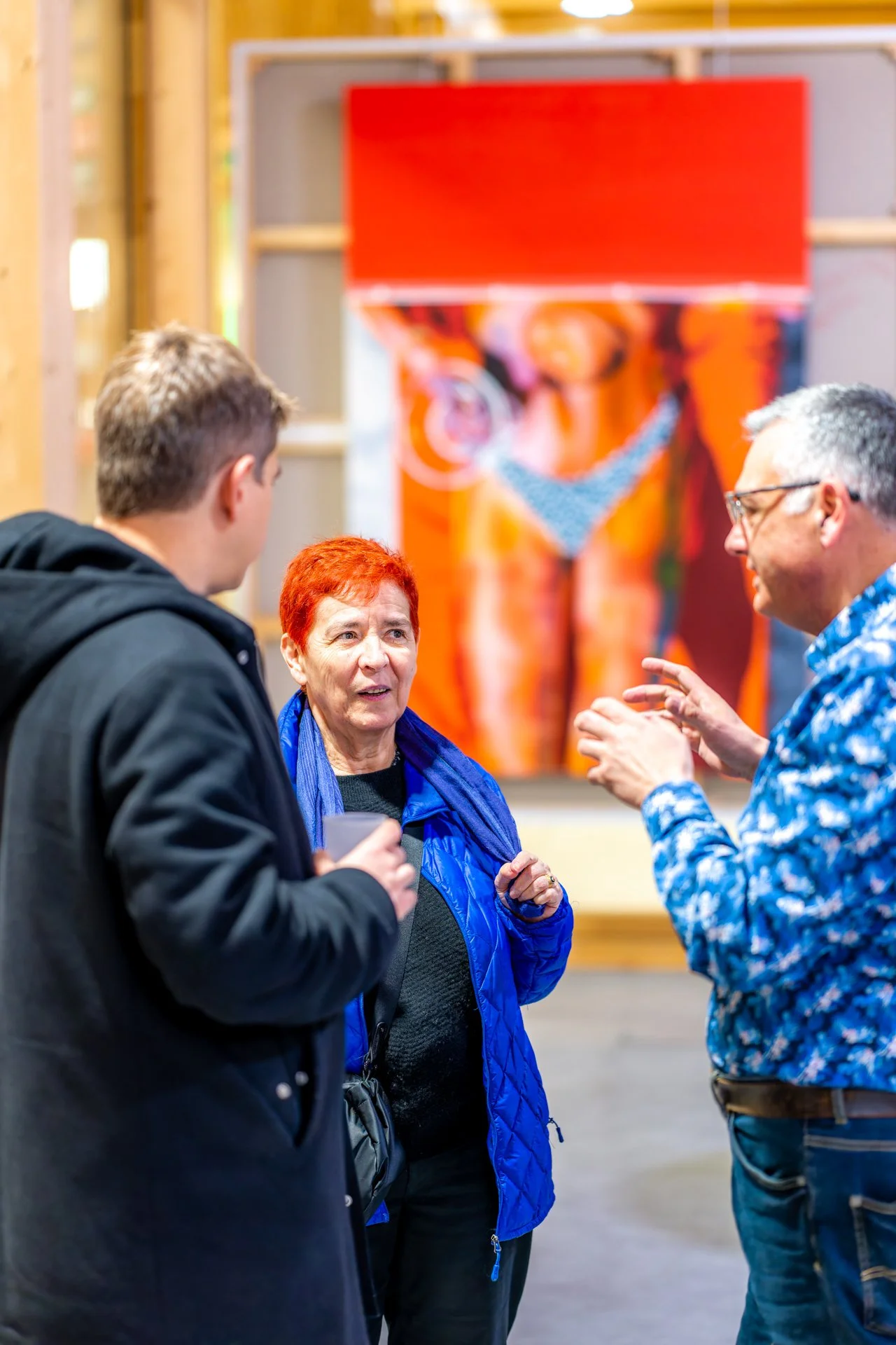 Trois personnes discutent dans une galerie d'art avec une grande peinture abstraite en arrière-plan.