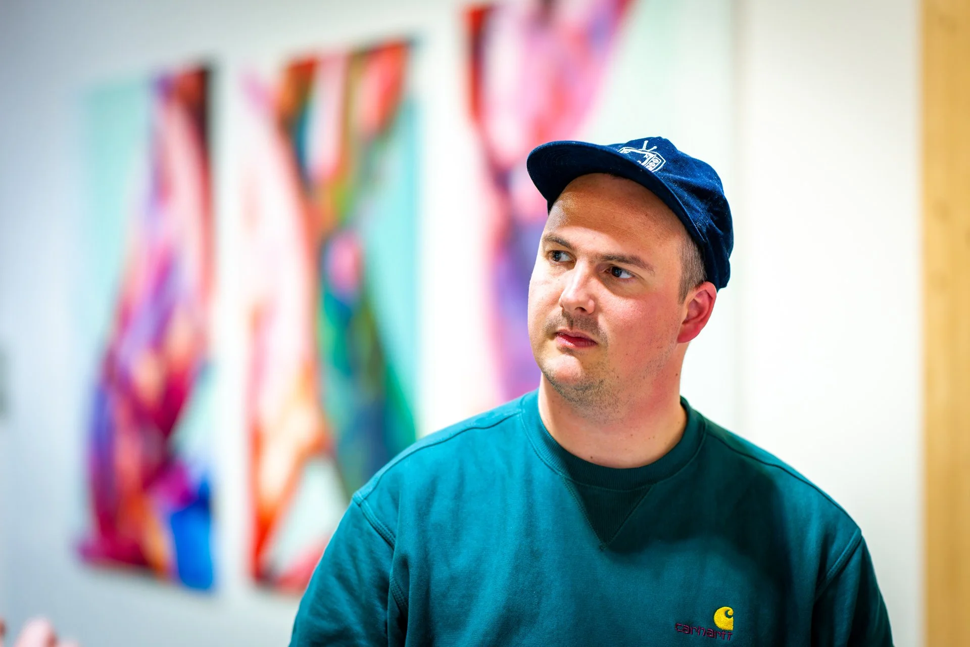 Un homme jeune portant une casquette bleue et un sweatshirt vert foncé, se tenant dans une galerie d'art avec des œuvres colorées floues en arrière-plan.