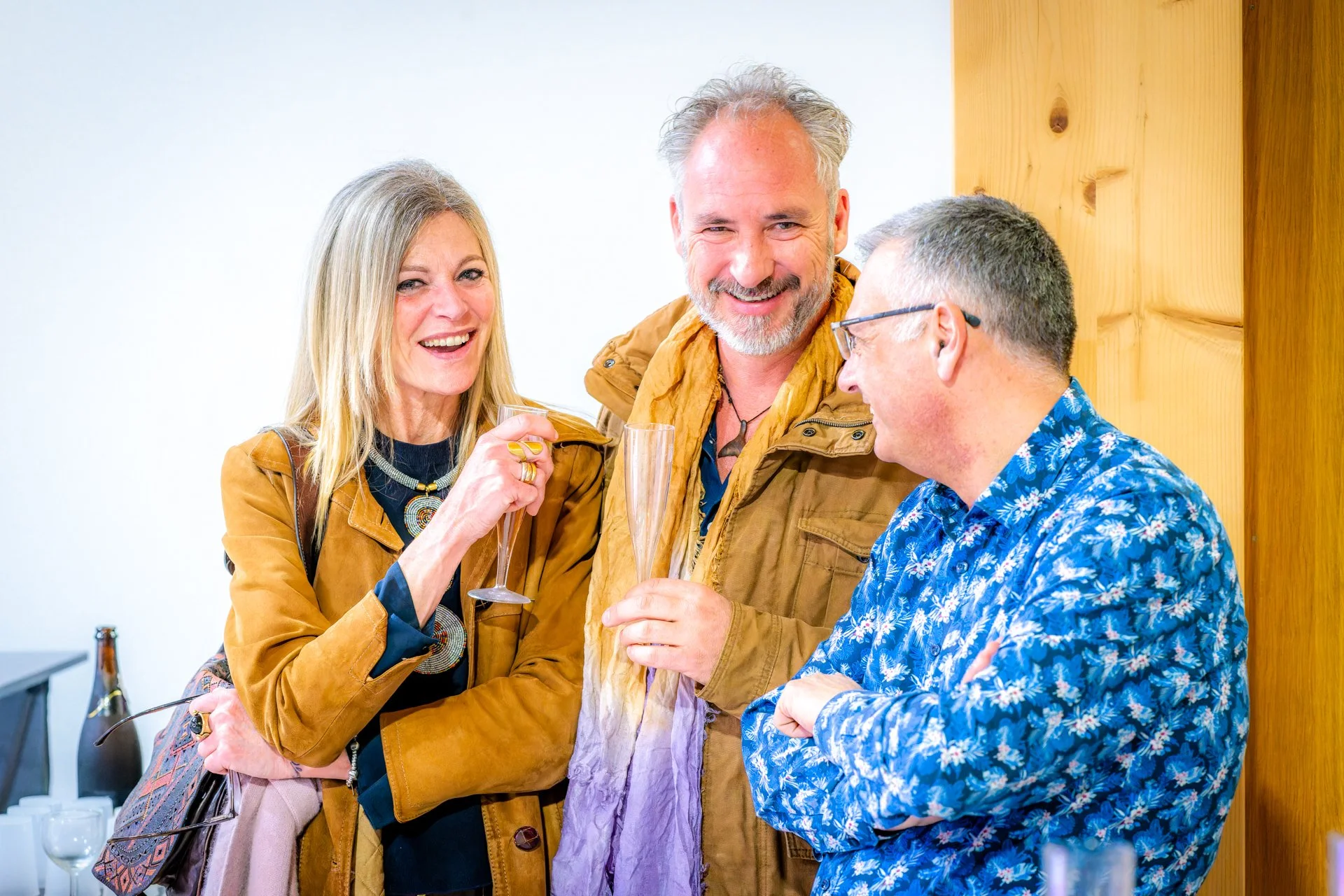 Trois personnes souriantes discutent avec des flûtes à champagne dans les mains. Une femme tient ses lunettes et un sac à main.