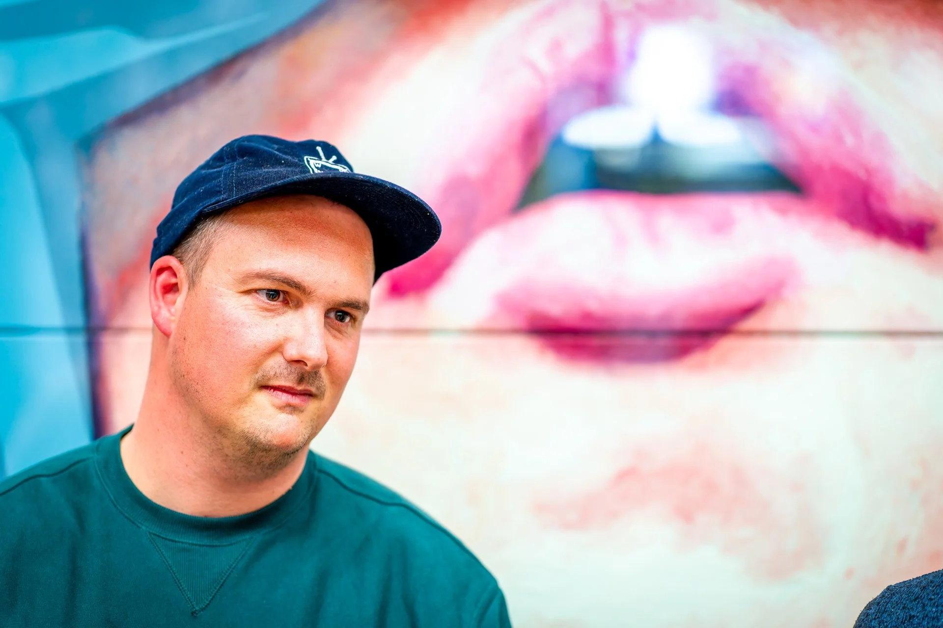 Homme portant une casquette, devant un mur coloré avec un portrait artistique de lèvres en arrière-plan.