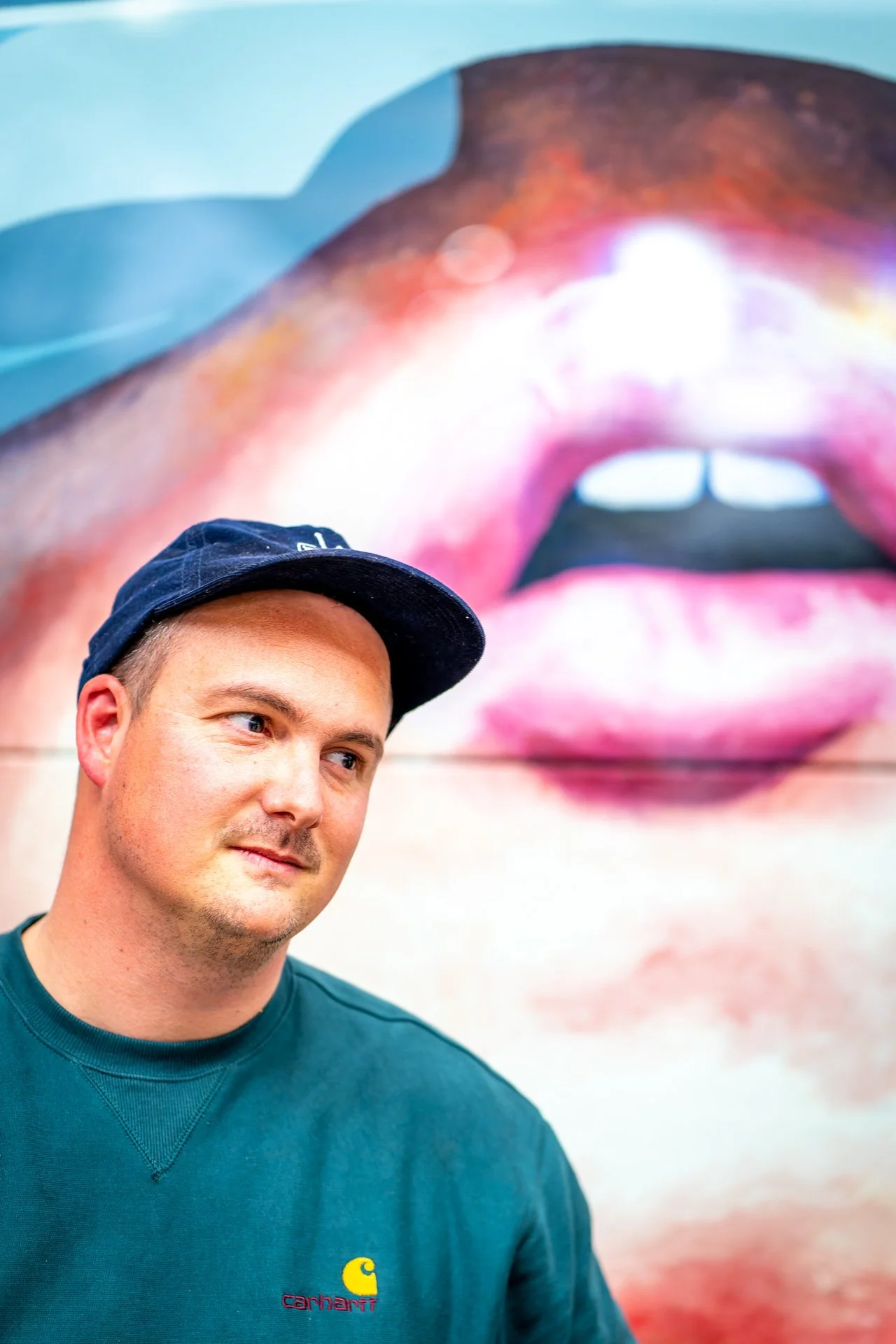 Homme portant une casquette bleue devant une peinture murale colorée représentant des lèvres.