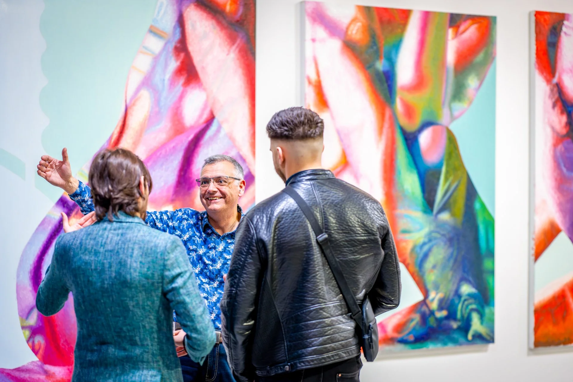 Trois personnes discutant devant des œuvres d'art colorées exposées dans une galerie.