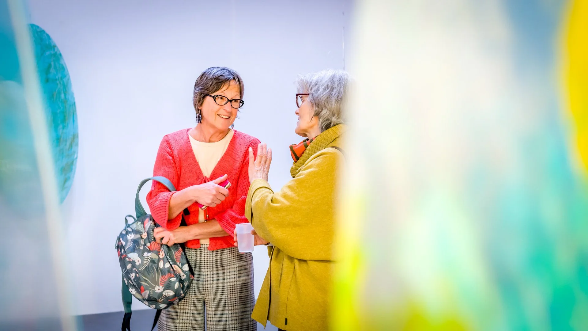 Deux femmes discutant dans une galerie d'art avec des œuvres colorées en arrière-plan.