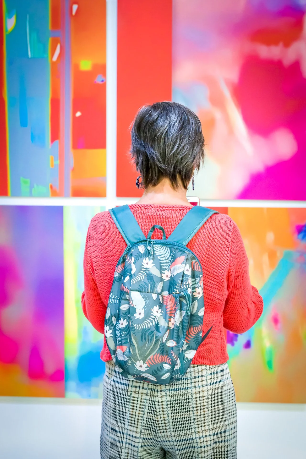 Personne de dos avec un pull rouge, un sac à dos à motifs floraux, regardant des œuvres d'art colorées sur un mur.