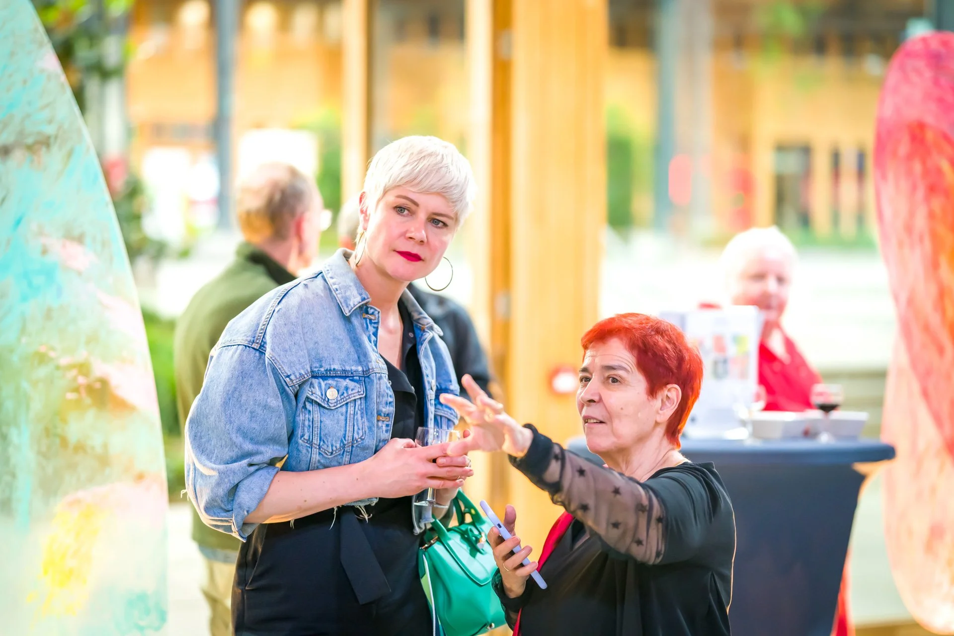 Deux femmes discutant dans une galerie d'art, l'une avec une veste en jean et l'autre avec une coupe de cheveux rouge, se tenant devant une sculpture colorée.