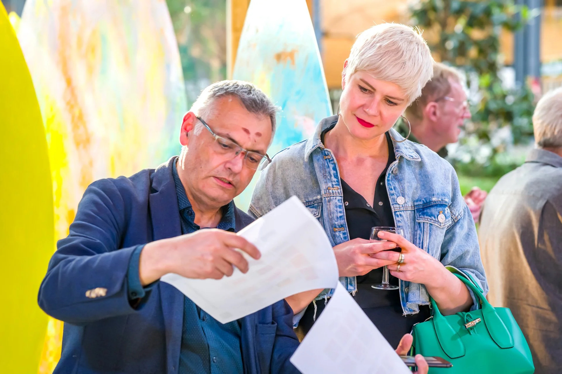 Deux personnes discutent en regardant des documents, l'homme tient les papiers tandis que la femme tient un verre de vin et un sac vert. Ils semblent être à un événement en plein air, avec d'autres personnes en arrière-plan.