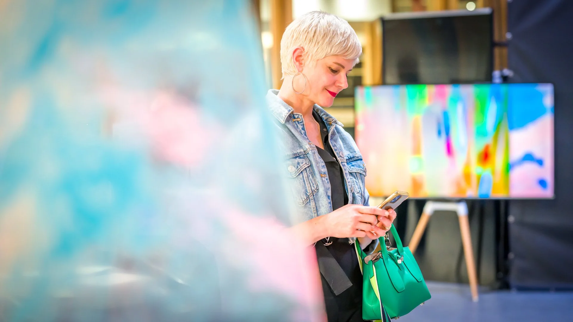 Une personne avec des cheveux courts tient un téléphone et regarde l'écran, portant une veste en jean et un sac vert vif. Arrière-plan coloré flou.