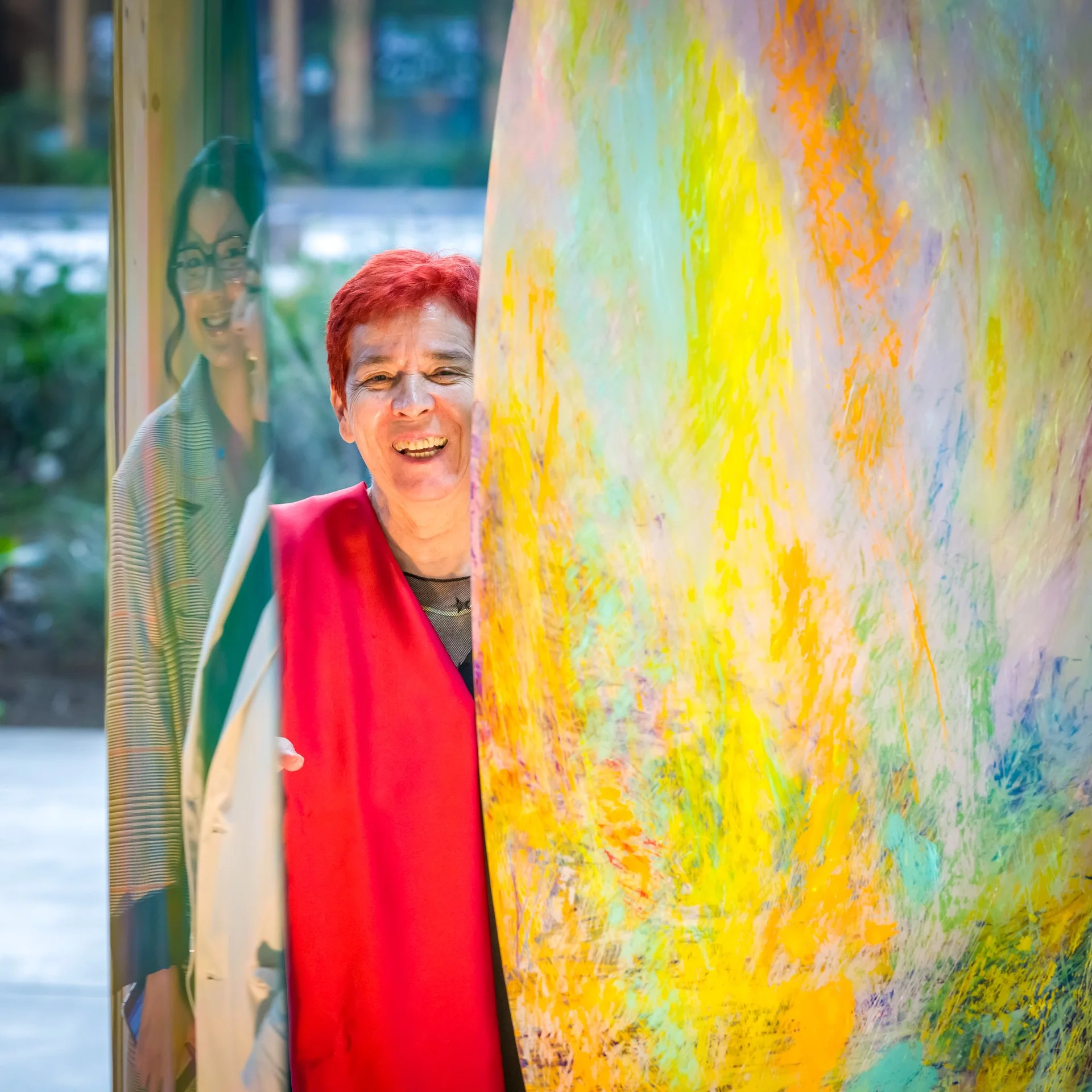 Une personne souriante se tient derrière une œuvre d'art colorée avec des motifs jaunes, bleus et violets. Elle porte un vêtement rouge. Au côté, un reflet d'une autre personne est visible dans une surface vitrée.
