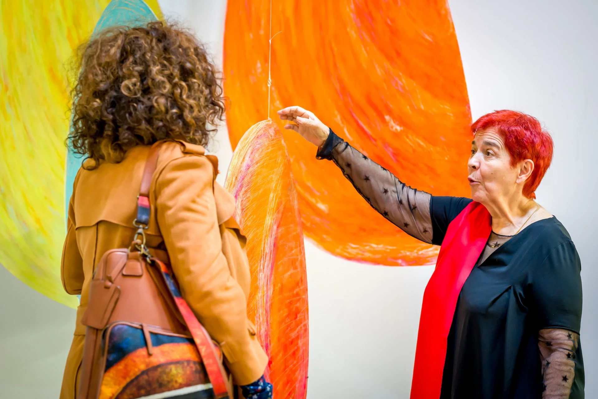 Photo pendant le cocktail de vernissage d'une exposition à Bruxelles. Deux femmes discutent devant des œuvres d'art colorées et suspendues dans une galerie.