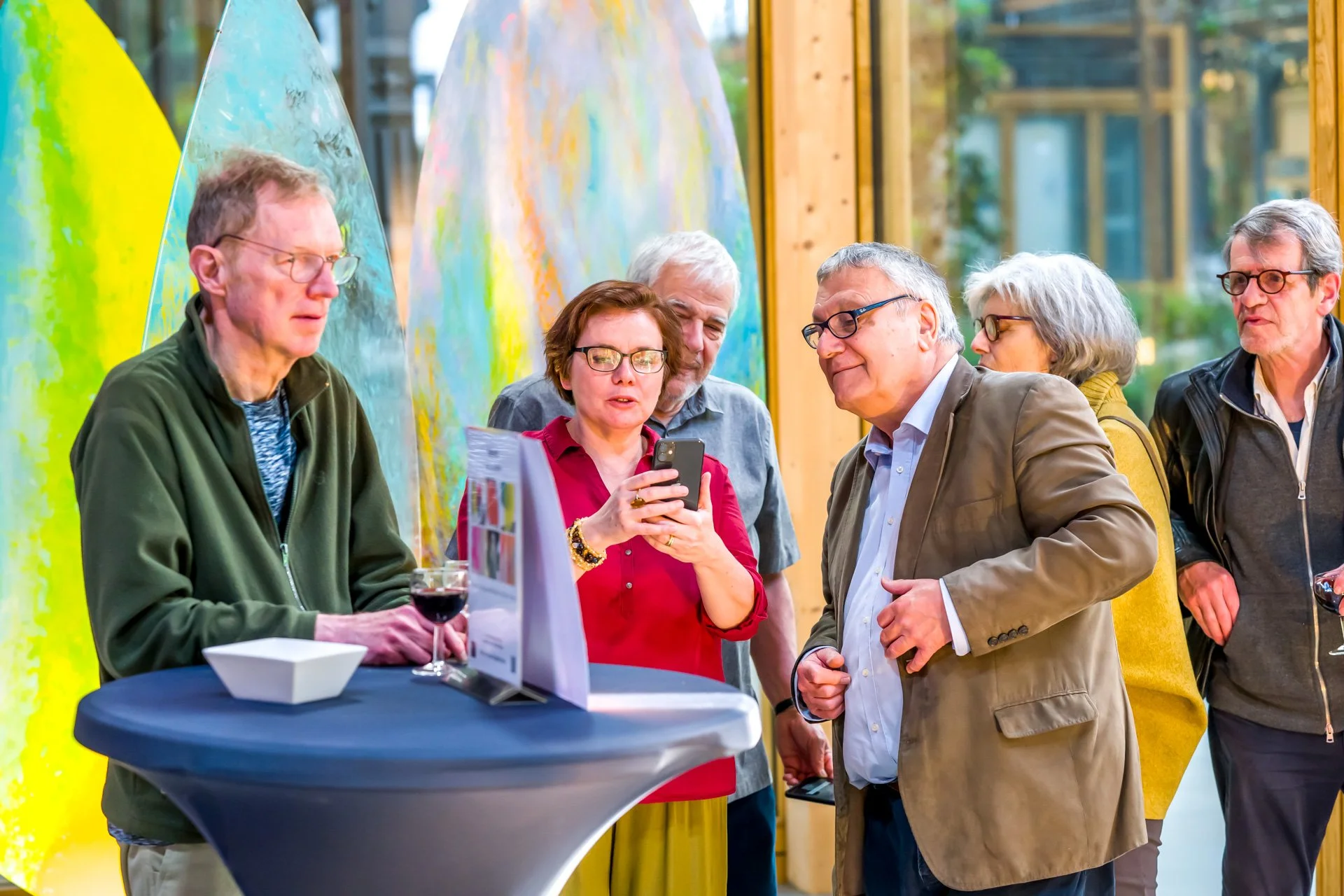 Un groupe de personnes âgées se tenant autour d'une table haute, regardant un smartphone. Décor coloré en arrière-plan.