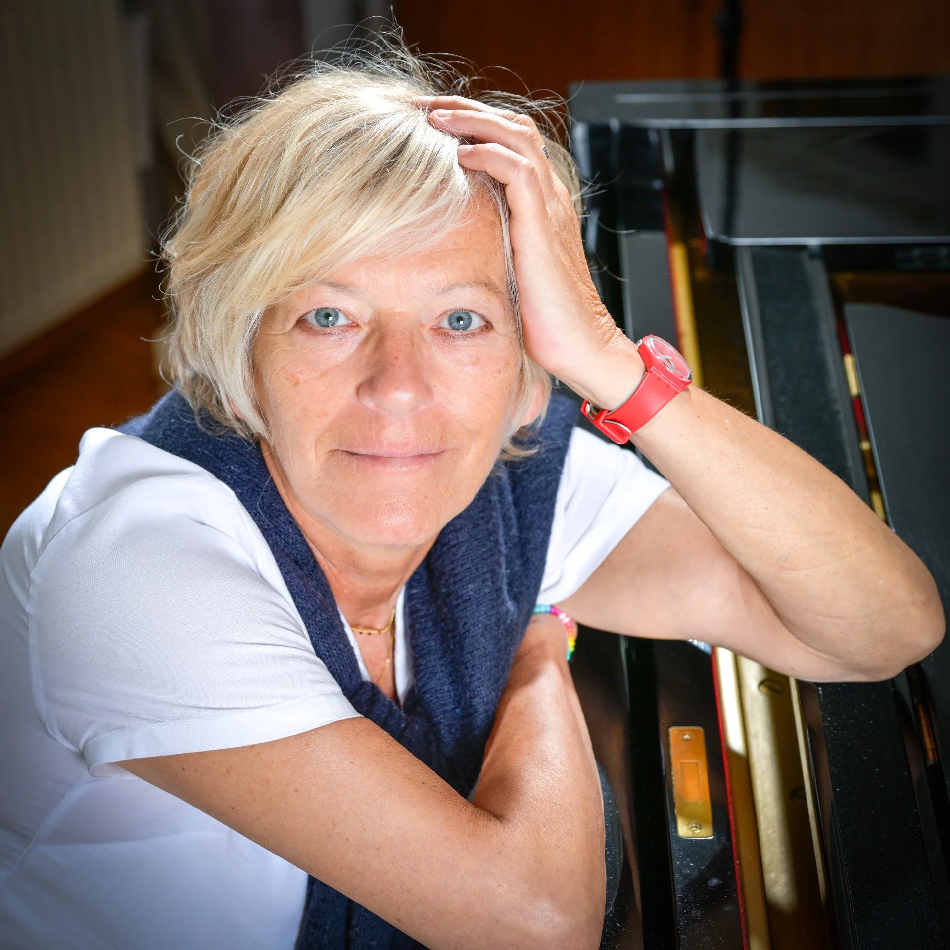 A woman with short blonde hair, wearing a red watch, leaning against a piano, looking pensive.