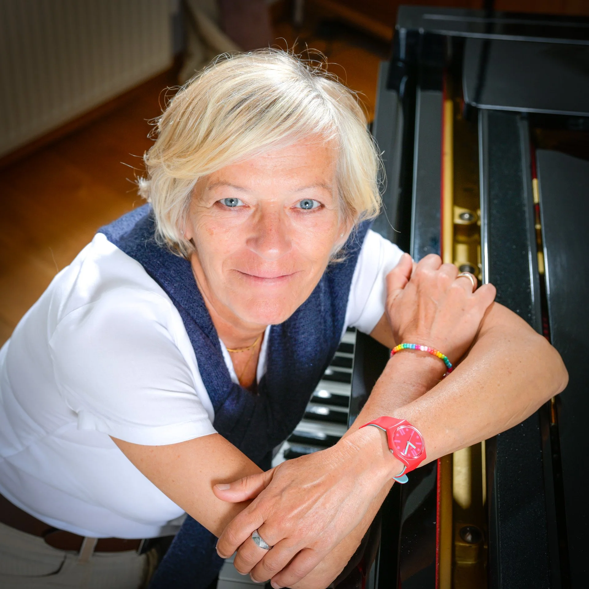 A smiling person with short blonde hair, posing next to a piano, wearing a white T-shirt and a multicoloured bracelet.