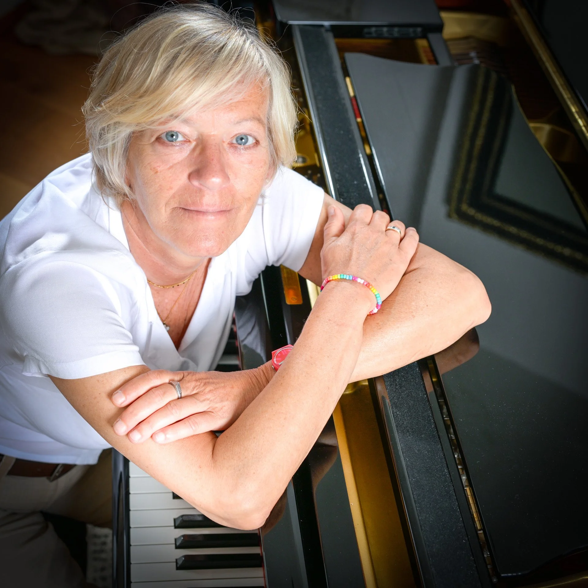 A person with blonde hair is sitting at a piano, with their arms crossed on the ledge, wearing a colourful bracelet.