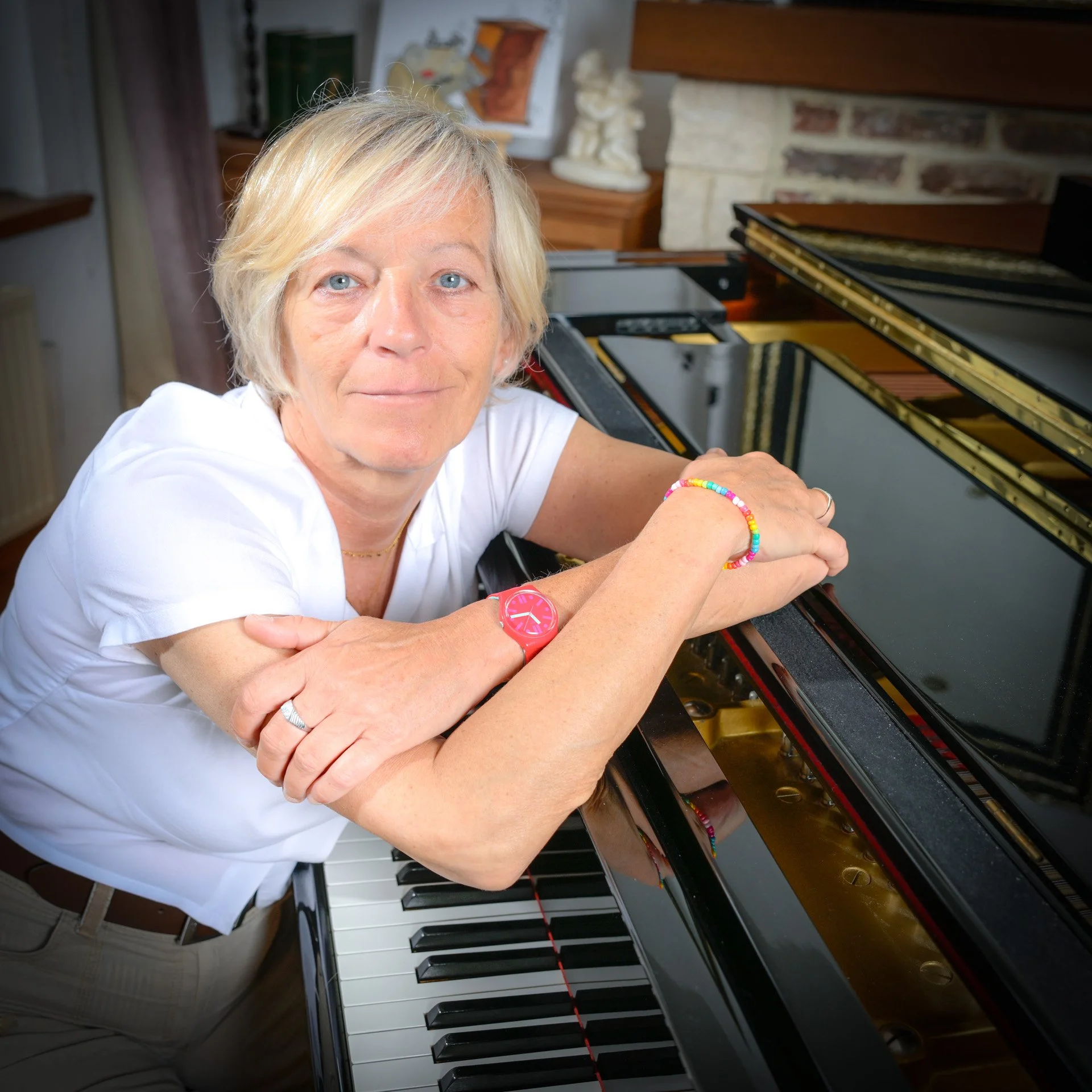 A person leaning against a grand piano, wearing a white shirt, a colourful bracelet and a pink watch, inside a room.