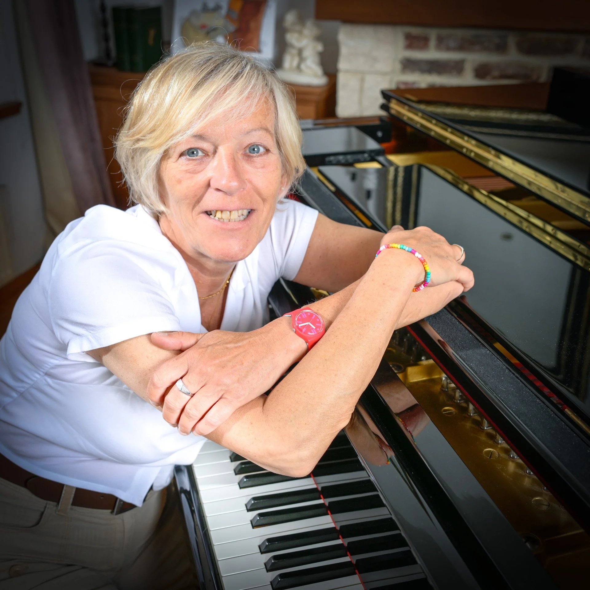 A person with blonde hair is smiling whilst leaning against a grand piano. They are wearing a pink watch and a colourful bracelet. The setting is probably an interior with a fireplace and decorative items in the background.