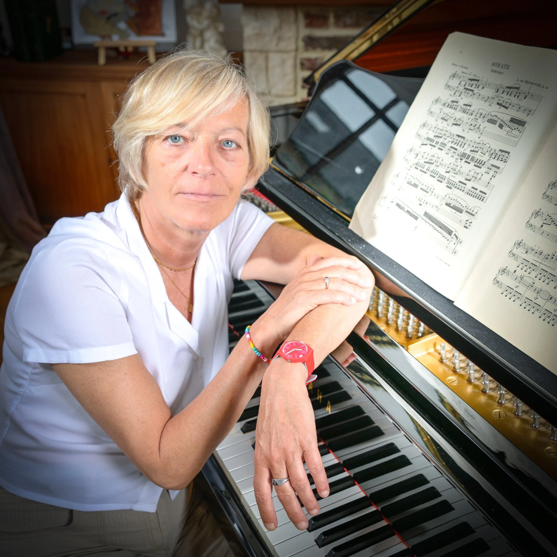 A blonde woman in a white shirt sitting on an upright piano, with an open sheet of music. She is wearing a red watch and a colourful bracelet."