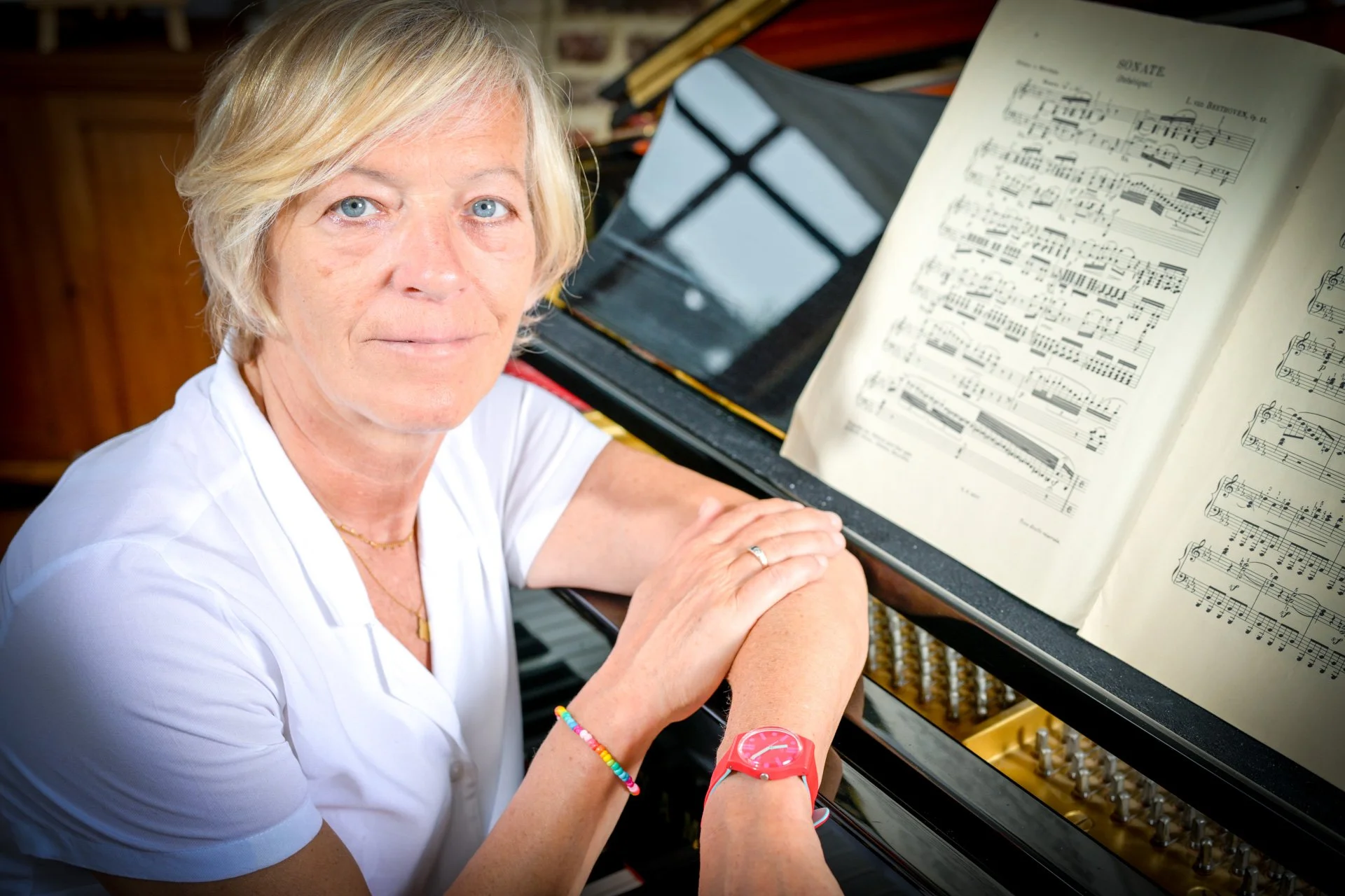A person sitting next to a piano with a sheet of music open in front of them. They are wearing a white shirt and a red watch.