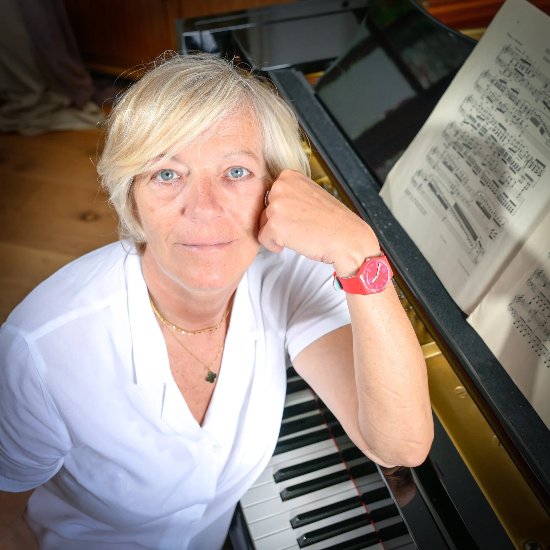 A person with blonde hair, wearing a white shirt, sitting next to a piano with sheet music open on it. She is wearing a red watch and appears to be looking upwards.