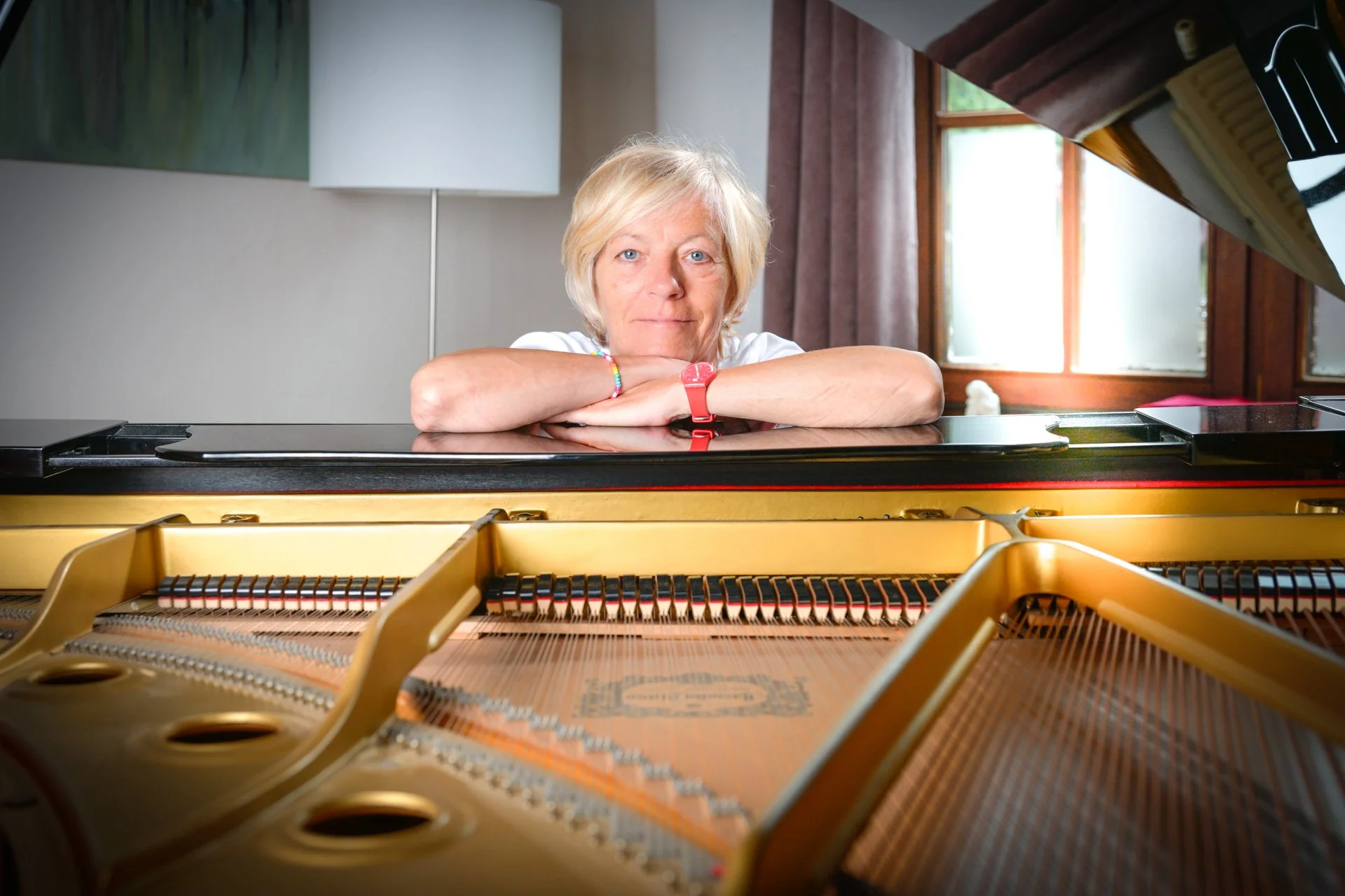 A blonde woman leaning against a grand piano, looking at the camera. The piano’s strings are visible inside.