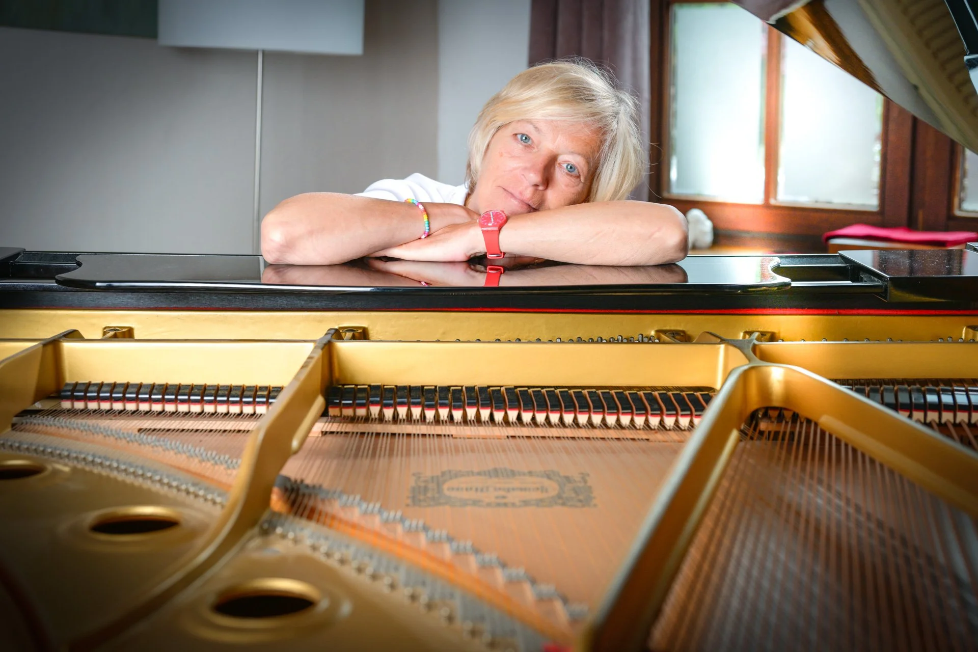 A blonde woman leaning against a grand piano, looking straight ahead, in a brightly lit interior.