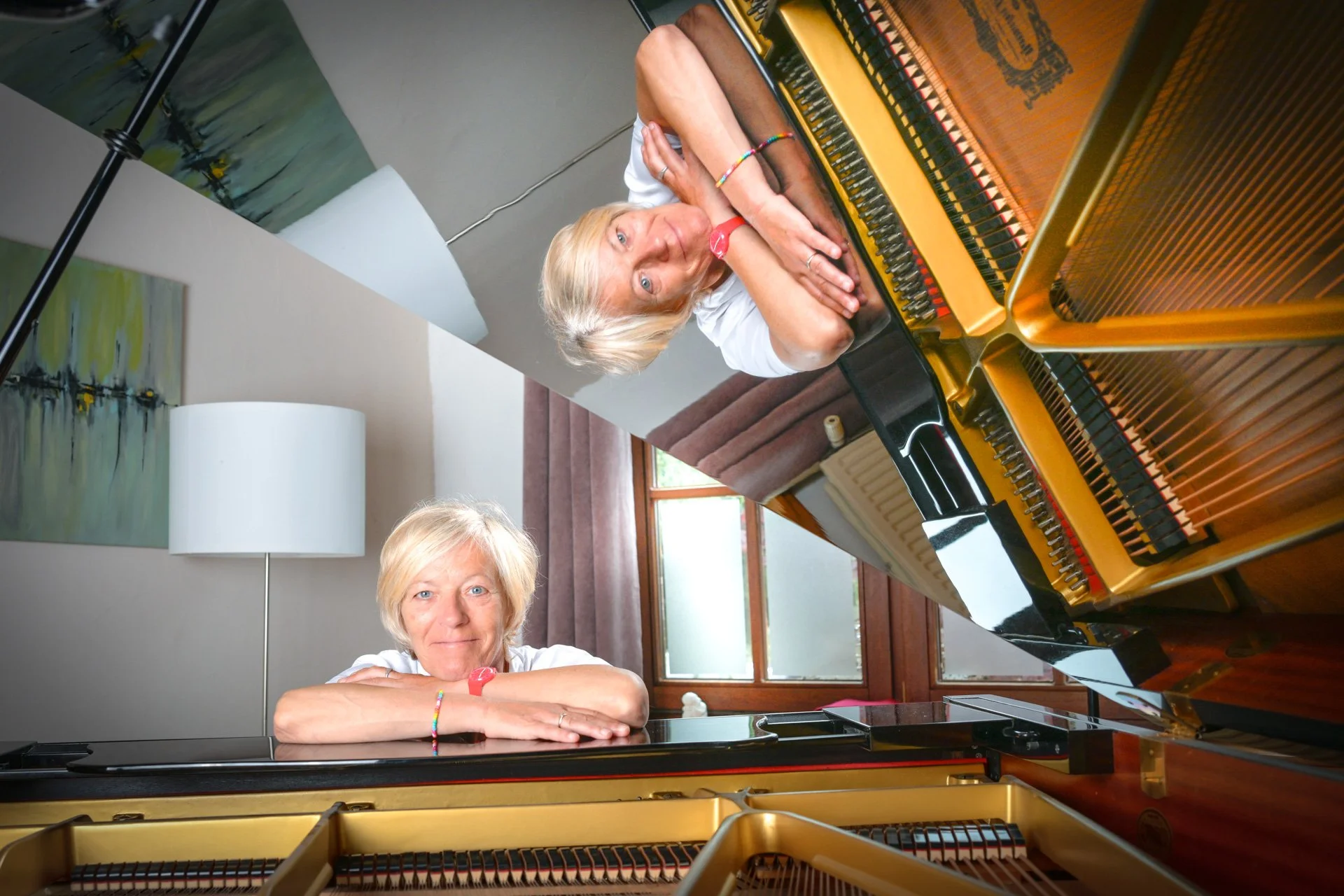 A woman with her arms crossed on a grand piano, her reflection visible in the piano lid. An interior scene featuring a window, a curtain, a lamp and a painting. The woman has short, grey hair and is wearing a colourful bracelet.