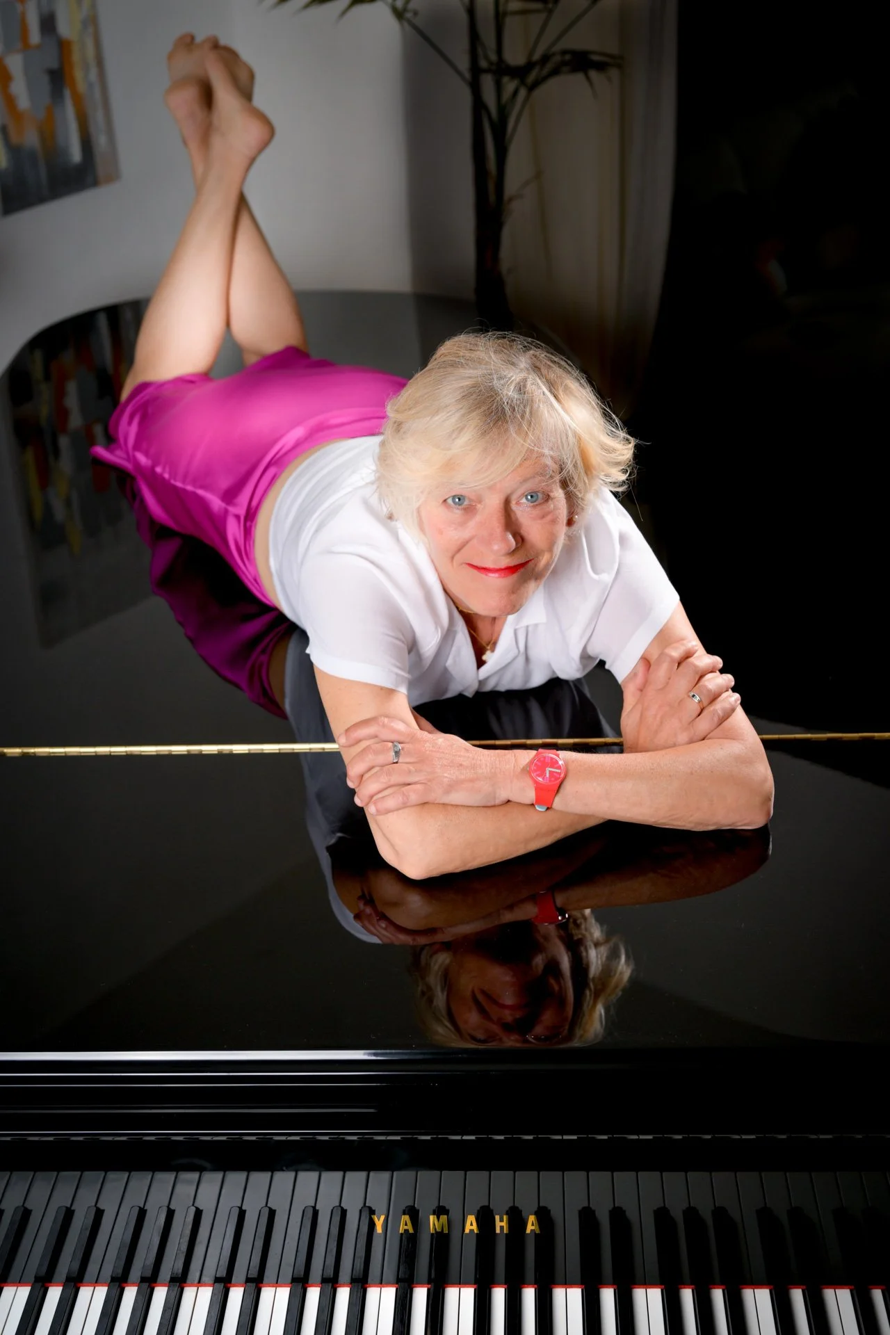 A woman lying on a Yamaha grand piano, wearing a pink skirt and a white shirt, with her reflection visible on the surface of the piano.