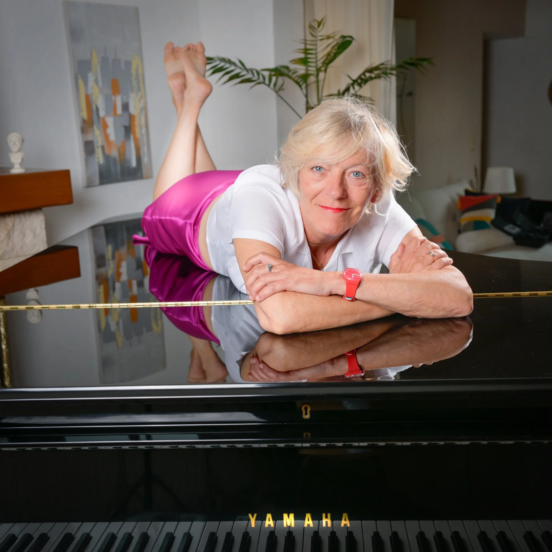 A woman lying on a Yamaha grand piano, wearing a pink skirt and a white blouse, smiling. An interior setting with plants and works of art in the background.