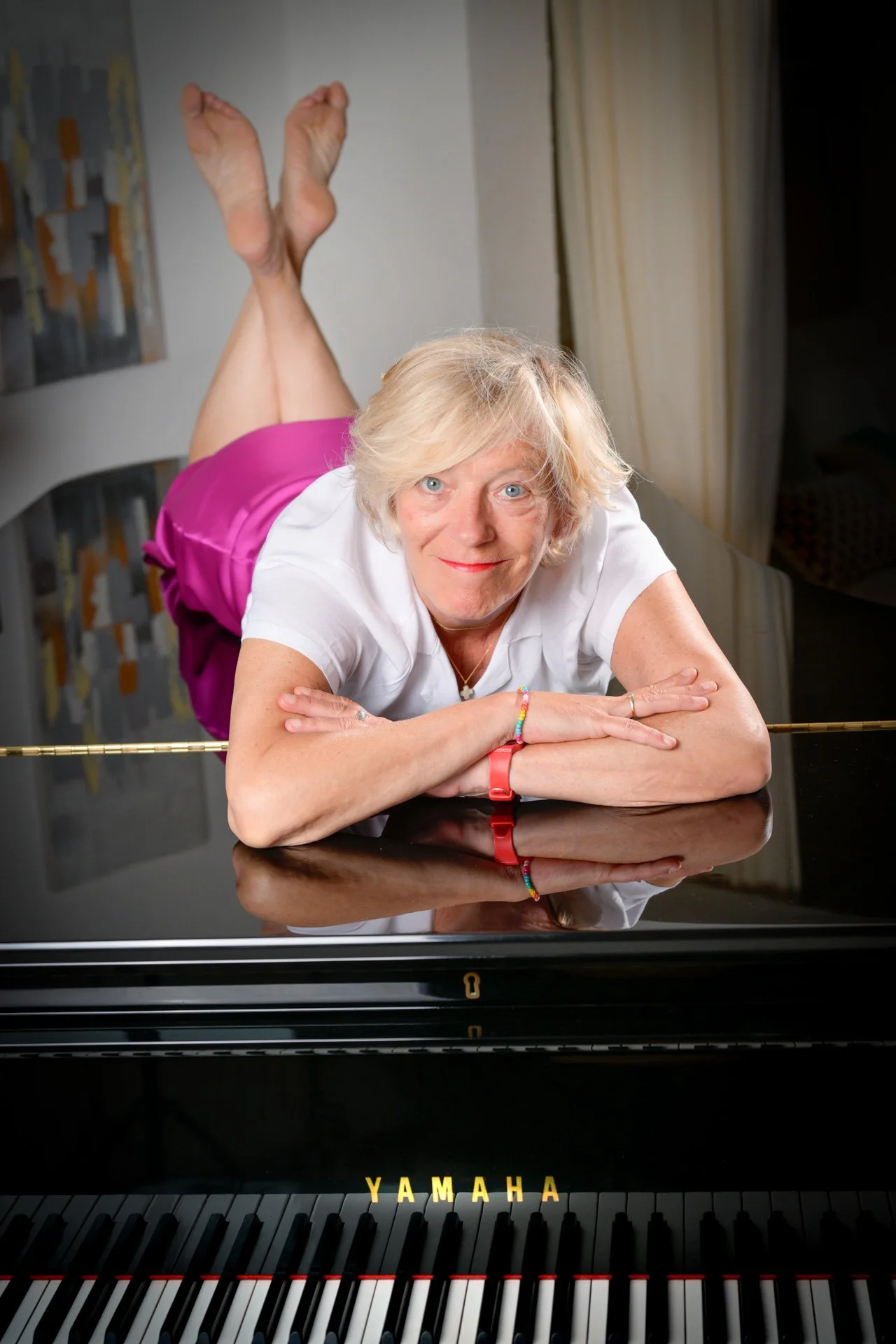 A woman lying on top of a Yamaha grand piano, with her feet in the air, wearing a white shirt and a pink skirt.