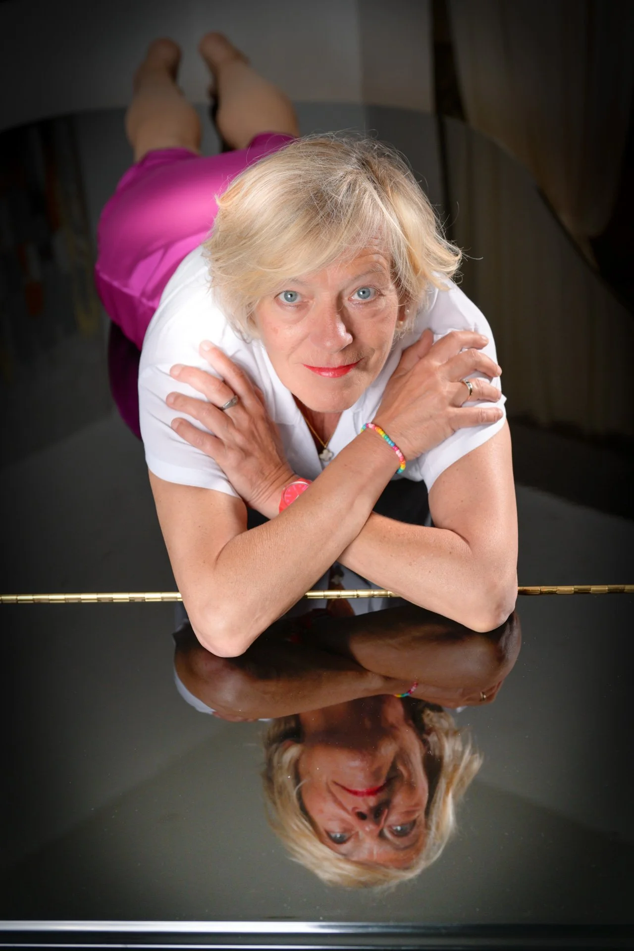 A blonde woman lying on a reflective surface, her arms crossed, wearing a pink dress and a colourful bracelet.