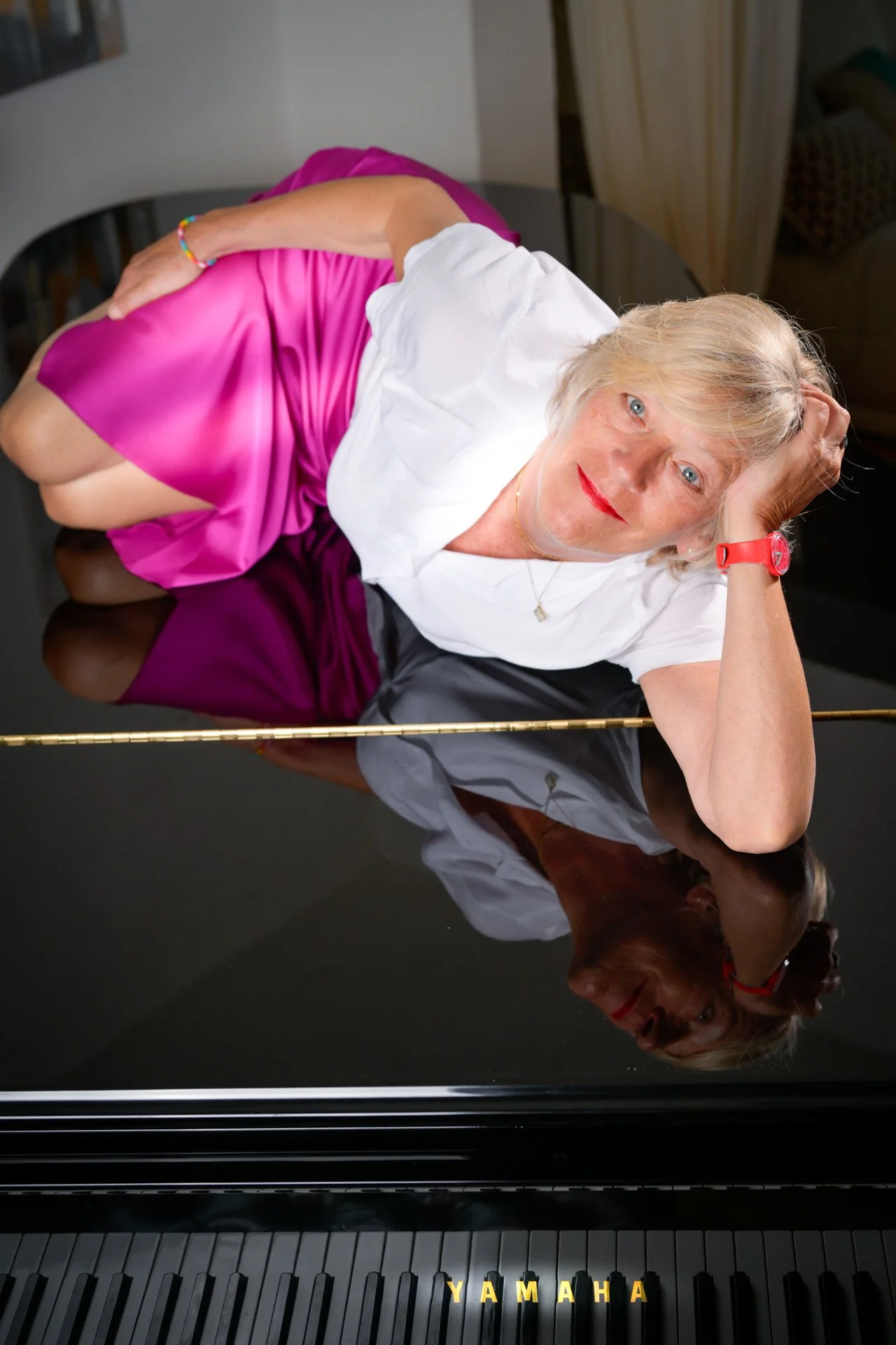 A woman lying on a black grand piano, wearing a pink skirt and a white blouse, looking towards the camera.
