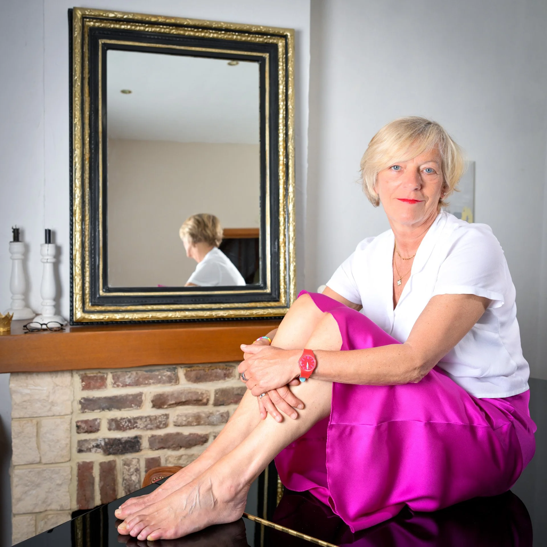 A woman sitting on a black table, wearing a white shirt and a pink skirt, with a mirror in the background reflecting her profile. An interior setting with an exposed brick wall.