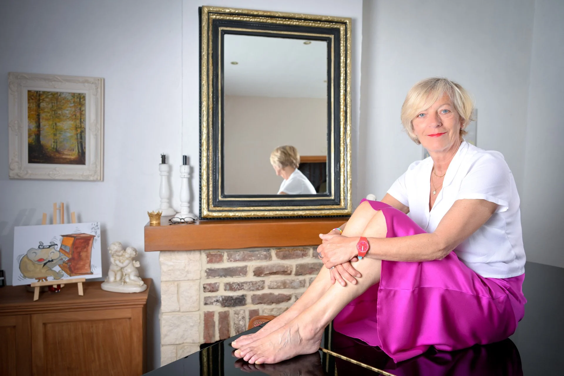 A woman sitting on a piano in a living room, wearing a pink skirt and a white shirt, with a mirror and wall decoration in the background.