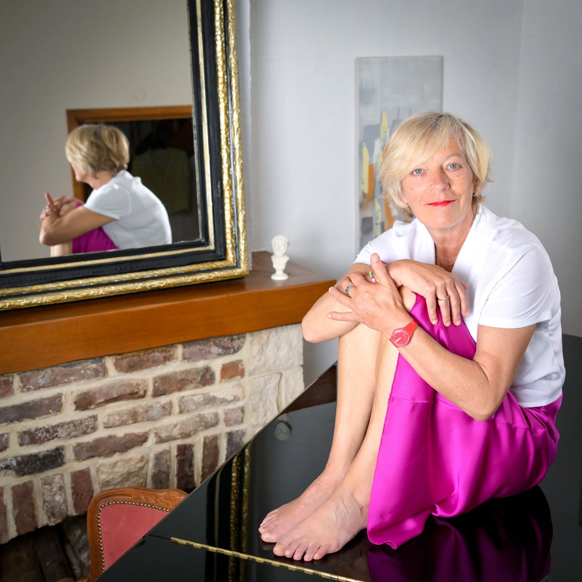 A woman is sitting on a black surface, wearing a pink skirt and a white top. She is standing in front of a mirror which reflects her image. The room features a stone fireplace and a gilded frame.