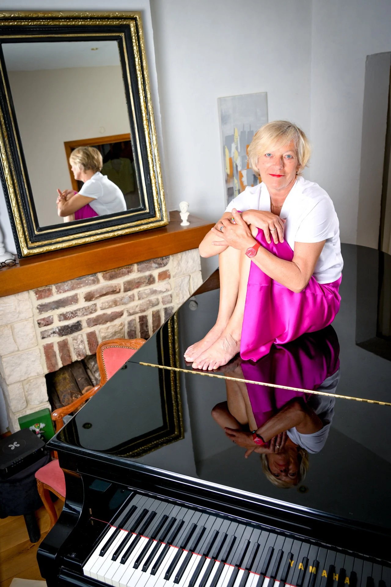 A woman sitting on a black grand piano, wearing a white top and a pink skirt, is reflected in the mirror behind her. A fireplace can be seen next to the piano.