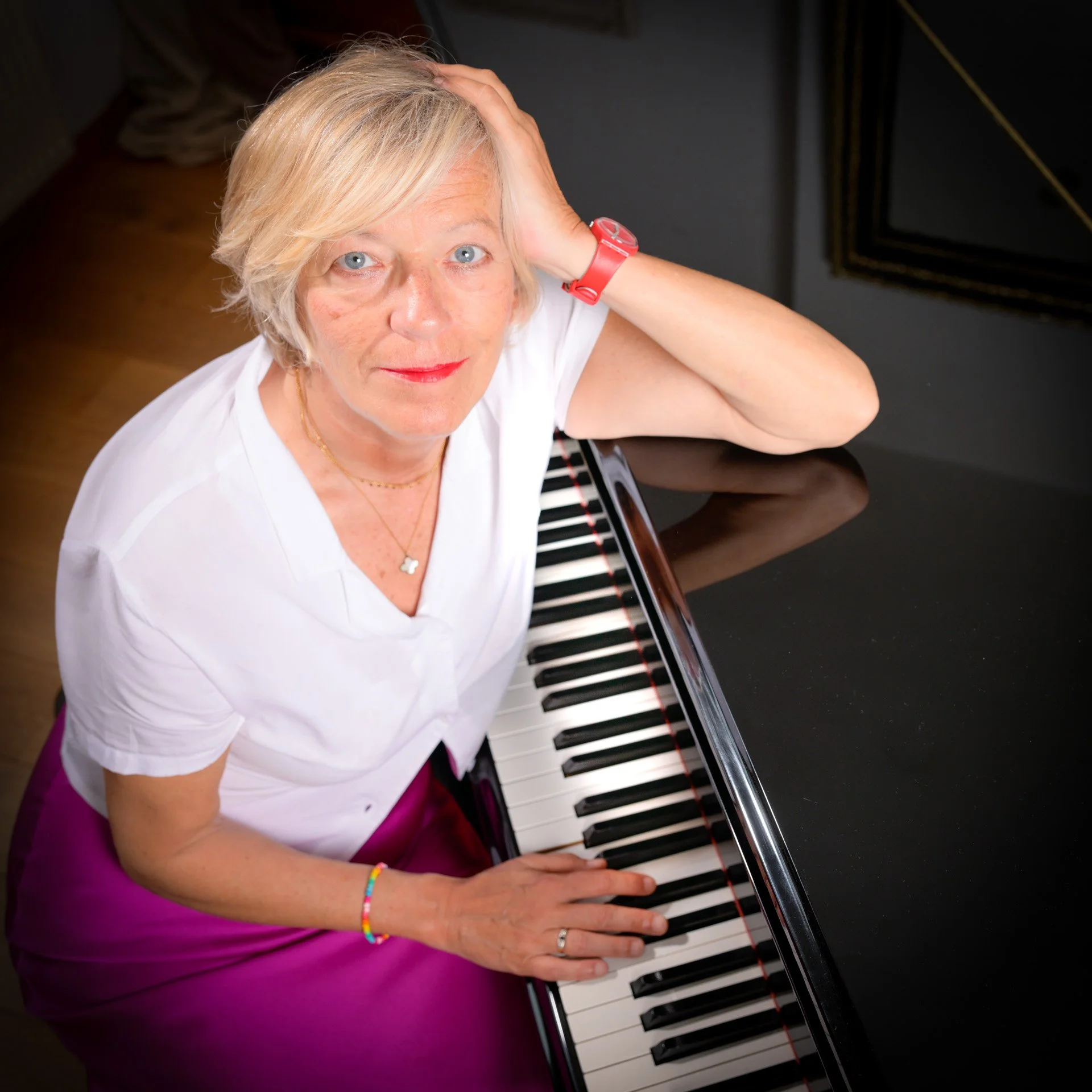 A woman with blonde hair and a white blouse is sitting at a piano, looking upwards. She is wearing a red watch and a pink skirt, with her hand resting on the piano keys.