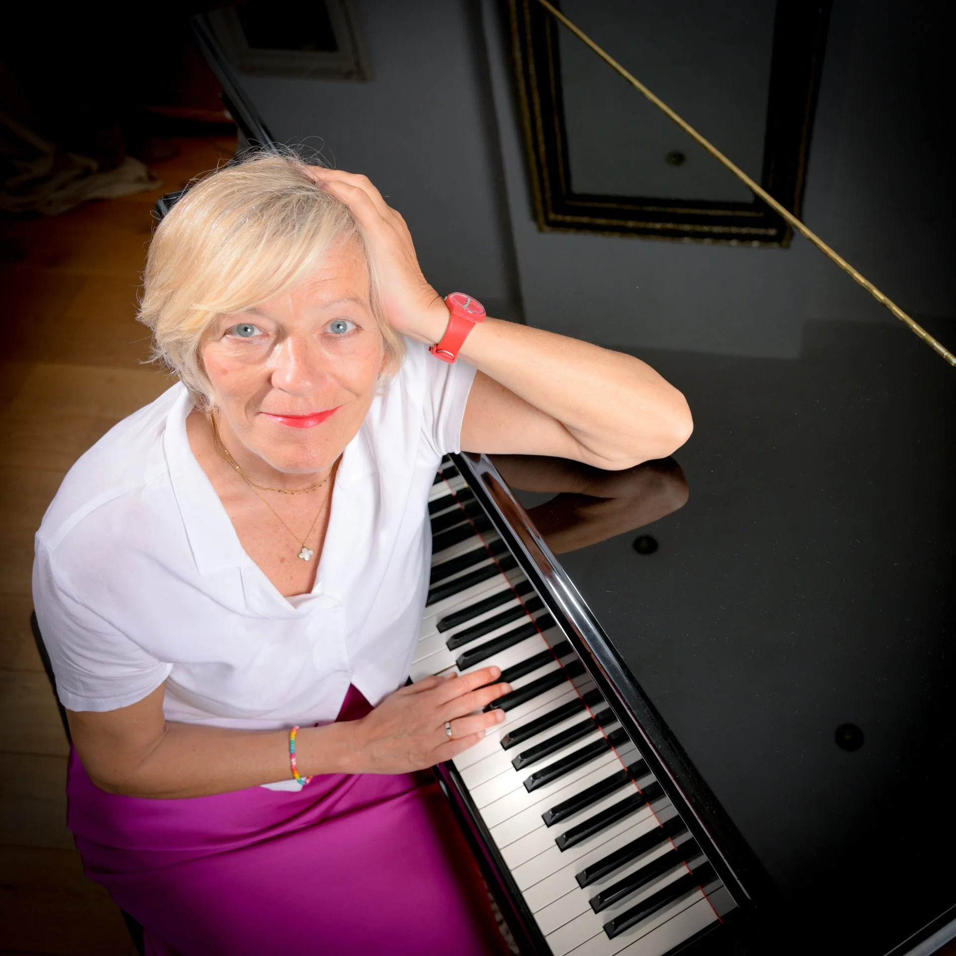 A woman with blonde hair posing next to a black grand piano, wearing a white blouse and a purple skirt.