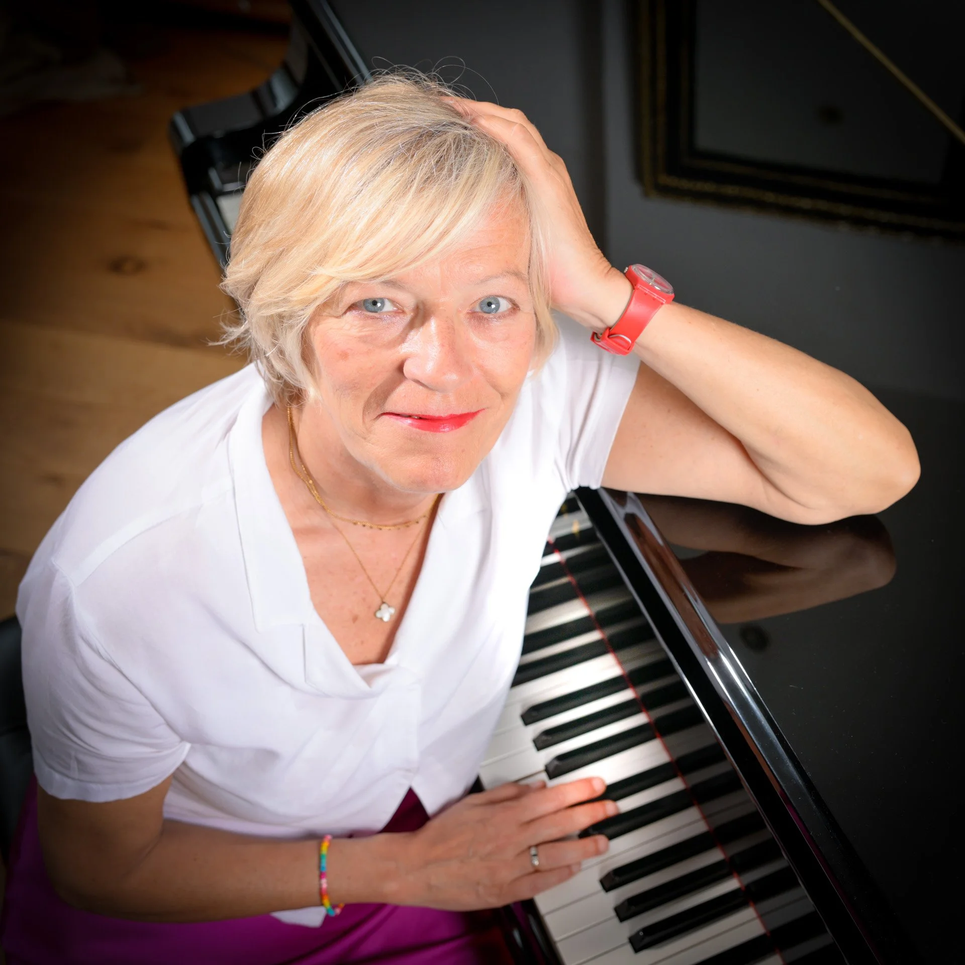 A smiling blonde woman sitting next to a piano, wearing a white shirt and a red bracelet. She is looking upwards, with one hand resting on her head.
