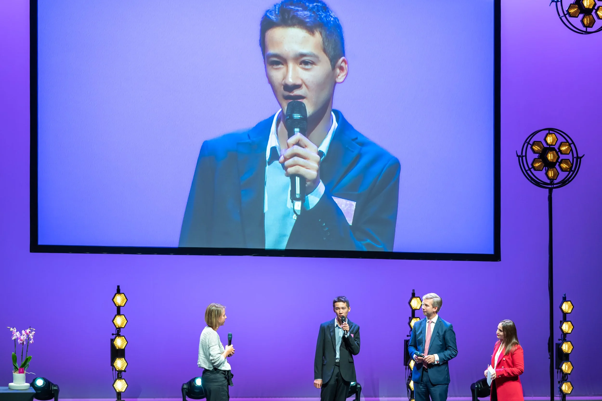 Un jeune homme en costume parle dans un microphone sur scène, projeté en grand sur un écran derrière lui, avec trois autres personnes à ses côtés, lors d'une conférence ou d'un événement formel, avec un décor violet et des lampes décoratives.