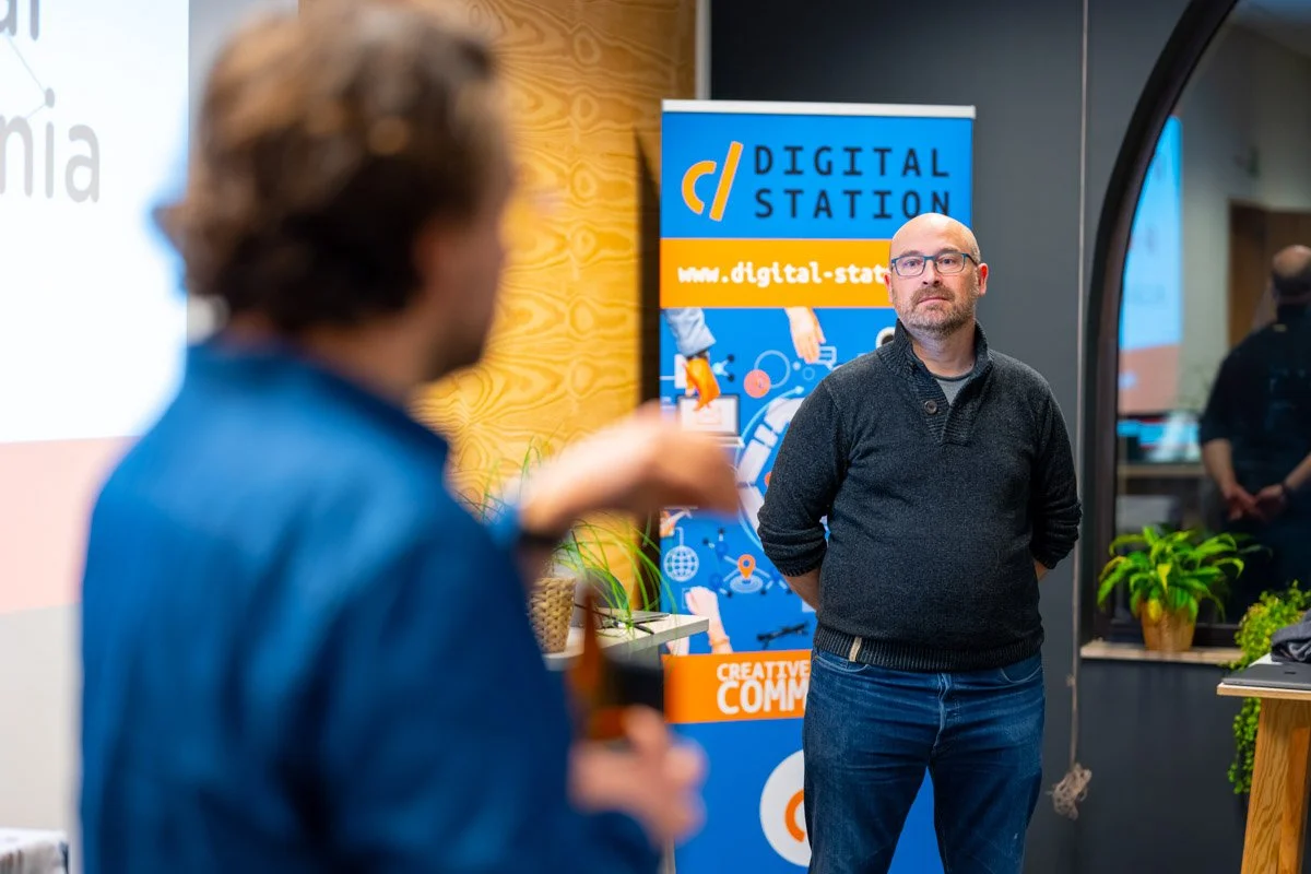 Denys Bornauw écoute un participant au premier plan, près d’une bannière Digital Station, lors du meetup WalChain à Charleroi.