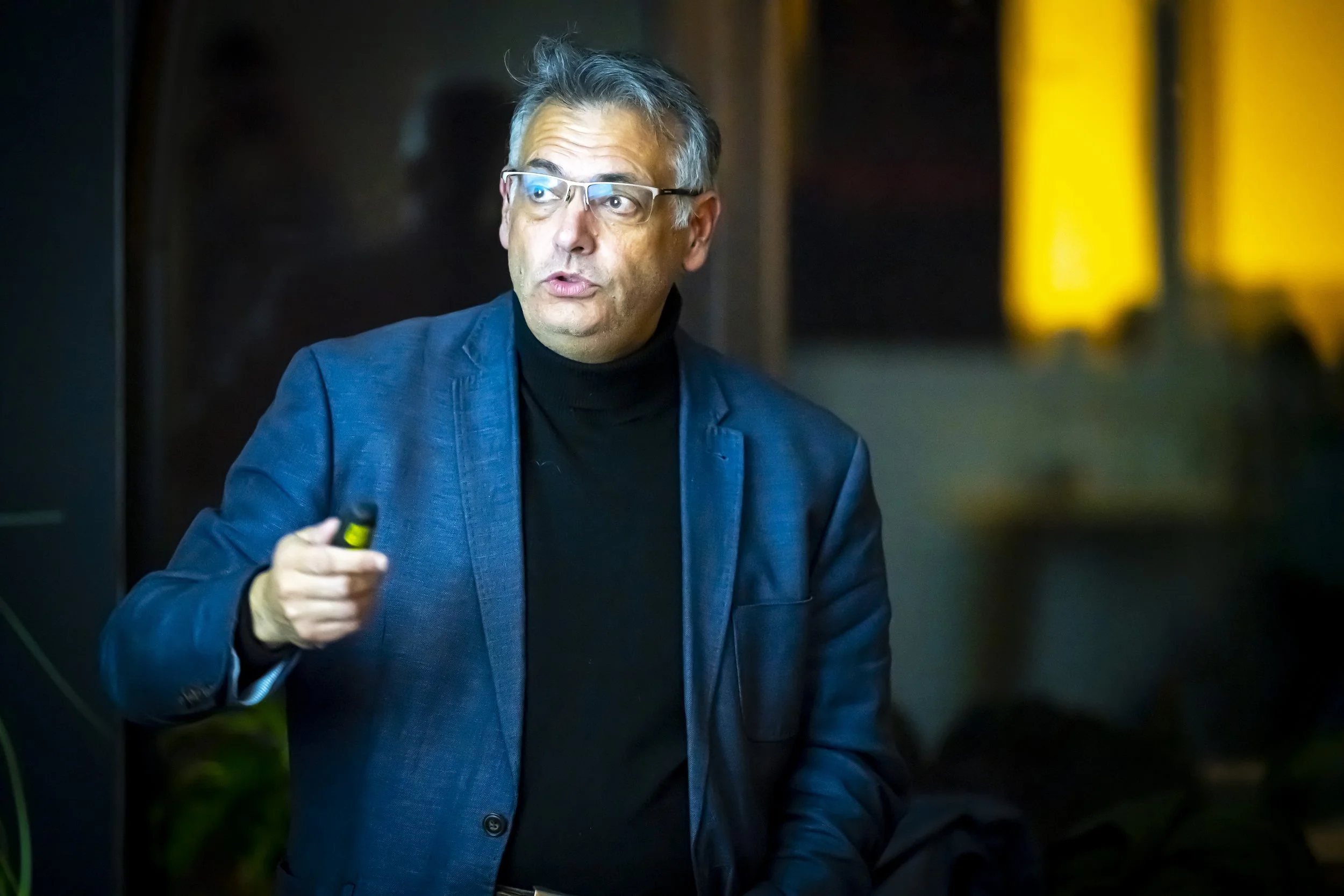 Un homme en costume bleu parle et tient un objet dans sa main, probablement lors d'une présentation ou conférence. Il porte des lunettes et semble concentré. L'arrière-plan est flou avec une lumière jaune.