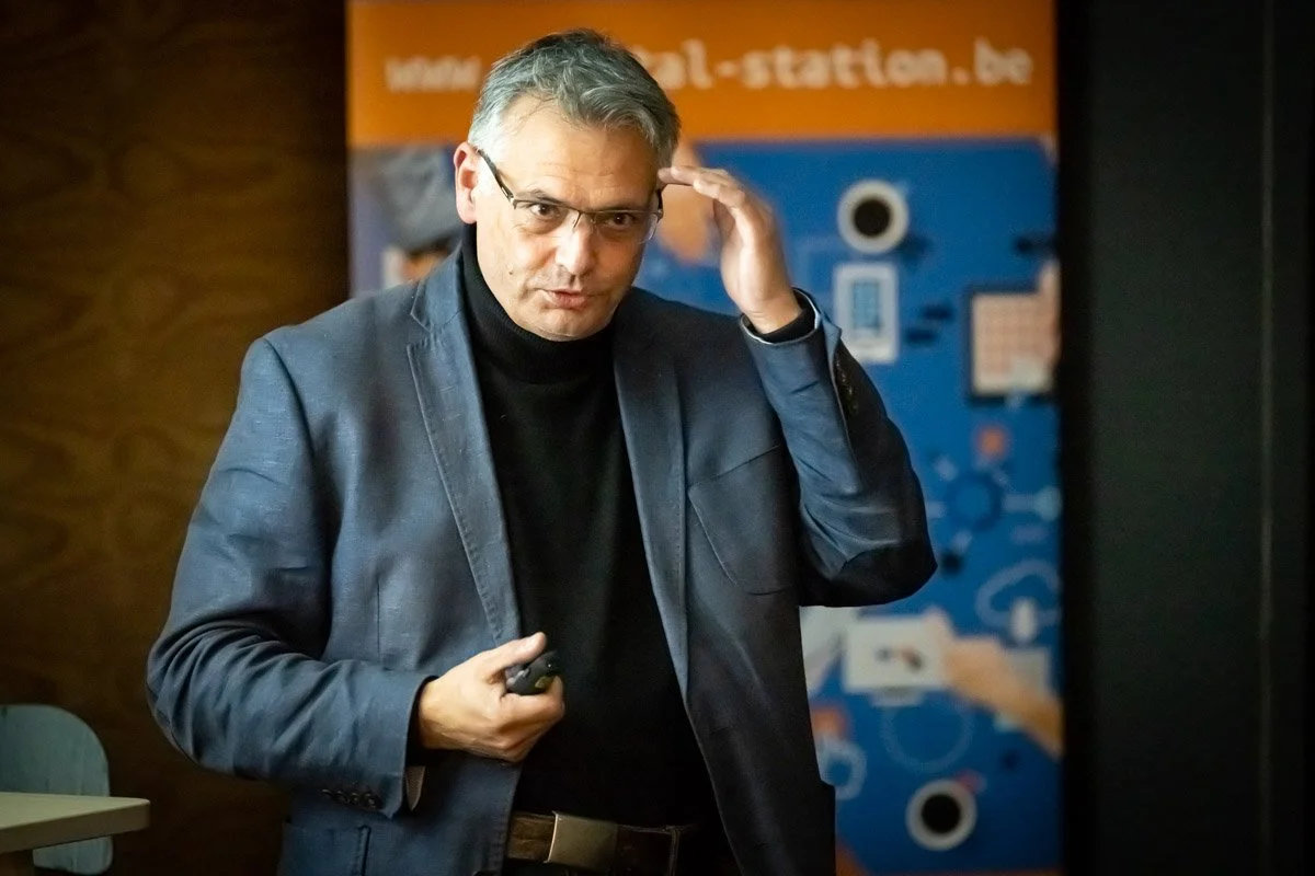 Vincent Parissis, en veste bleue et col roulé noir, porte une main à ses lunettes lors du meetup WalChain à Charleroi.